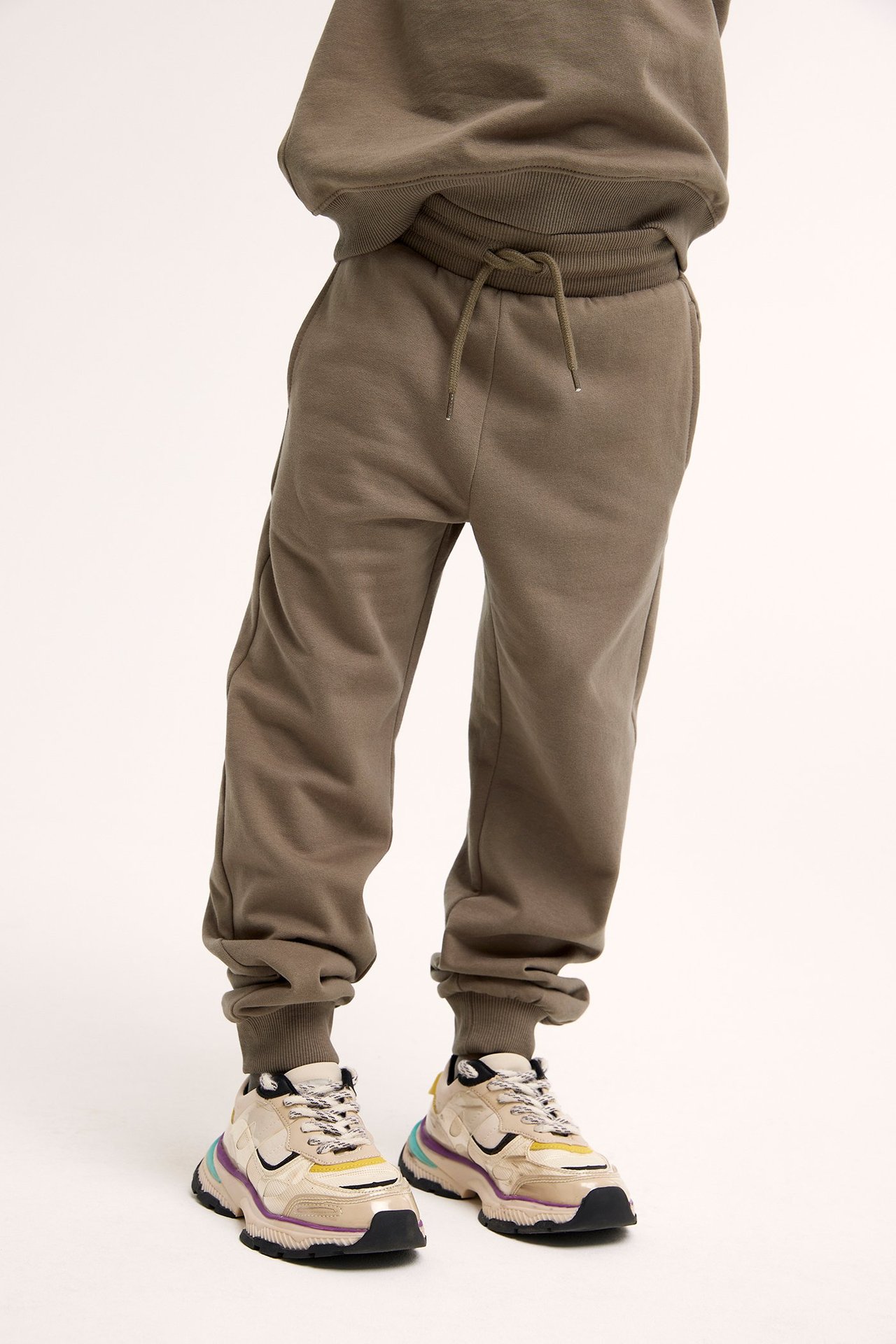 Edestäpäin: lapsi ruskeissa puuvillacollegehousuissa, resori ja kiristysnyöri, yhteensopiva pusero ja beiget lenkkarit.