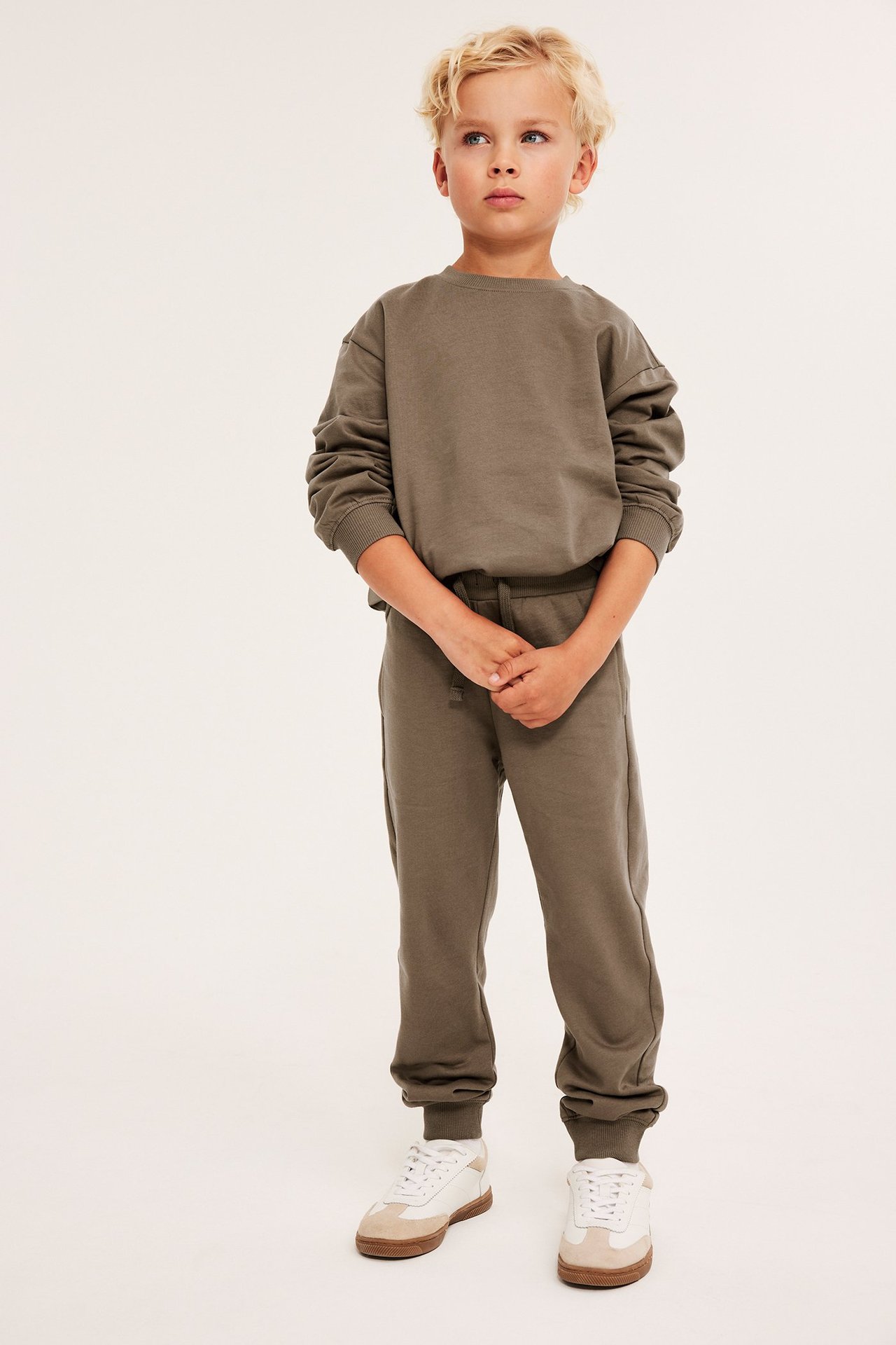 Widok z przodu dziecka w brązowych bawełnianych spodniach dresowych ze sznurkiem, pasującej bluzie i białych sneakersach.