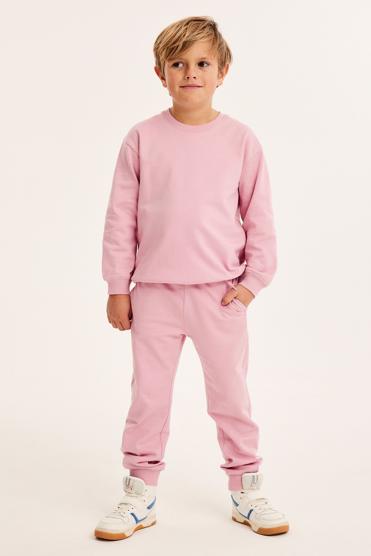 Frontvy av ett barn i ett ljusrosa jogging-set i bomull med muddar och vita sneakers.