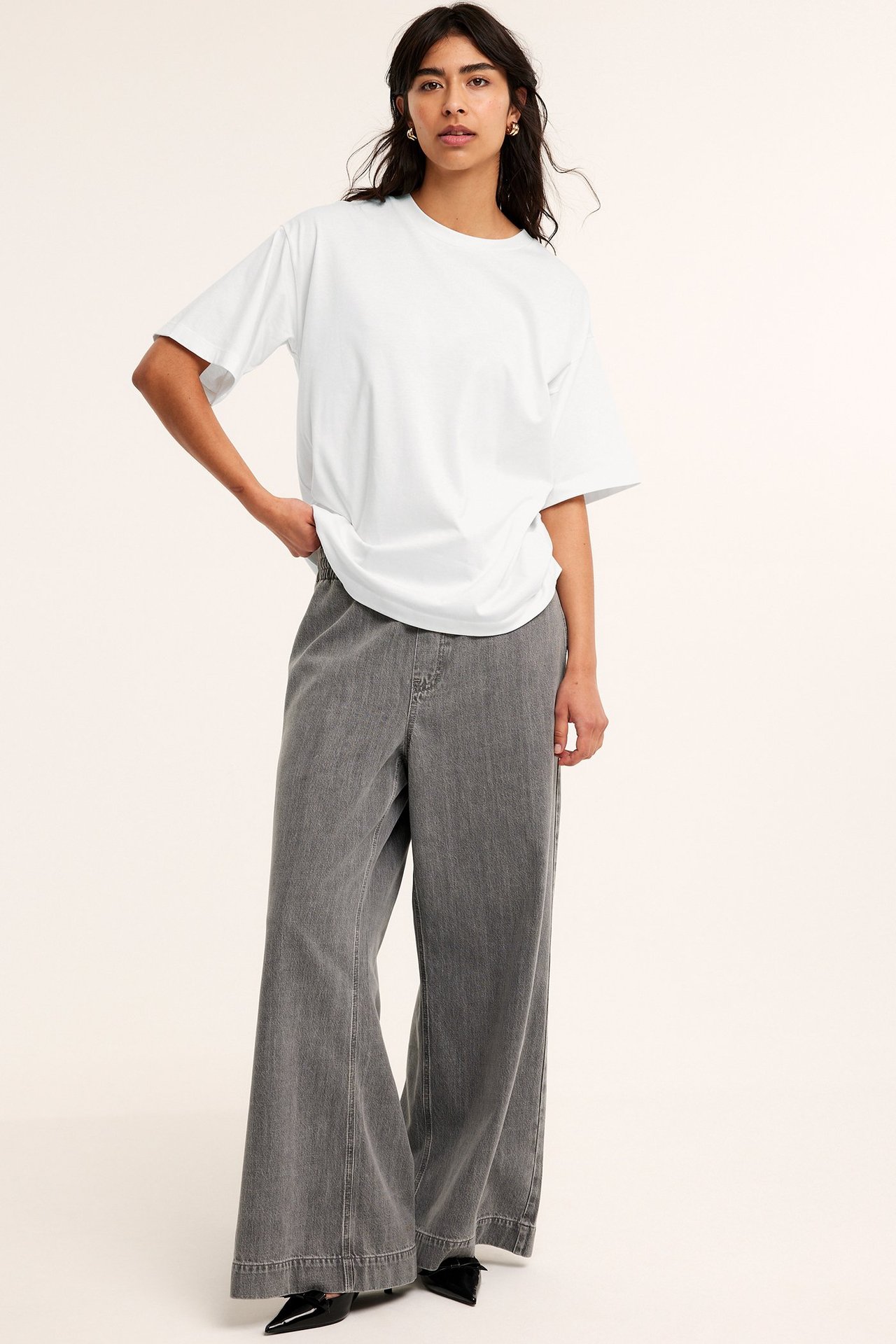 Widok z przodu: Kobieta w kremowym, oversize'owym bawełnianym T-shircie, szarych szerokich jeansach i czarnych butach.