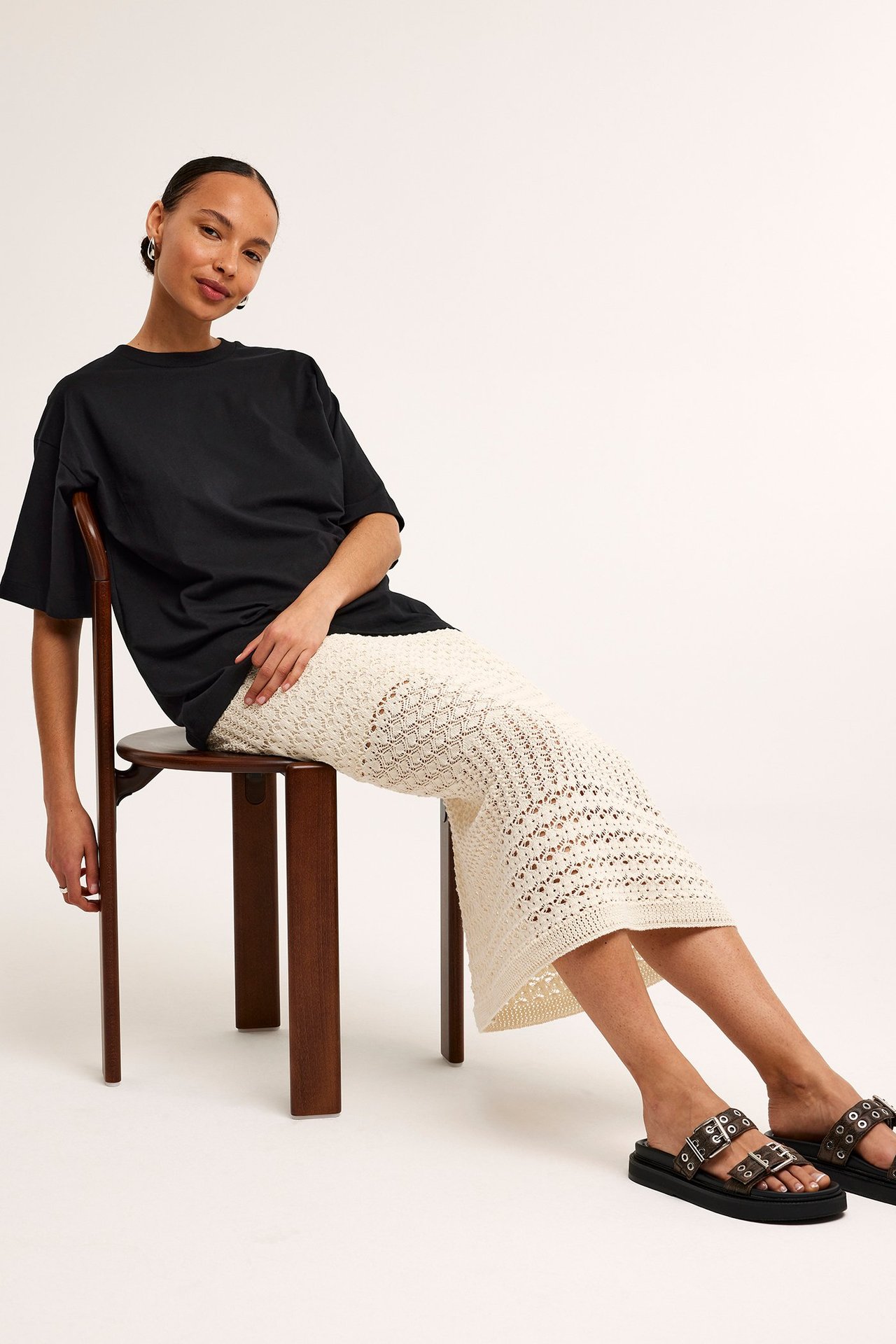 Forfra: kvinne i svart oversized bomulls-t-skjorte, kremfarget heklet midiskjørt og svarte platåsandaler.