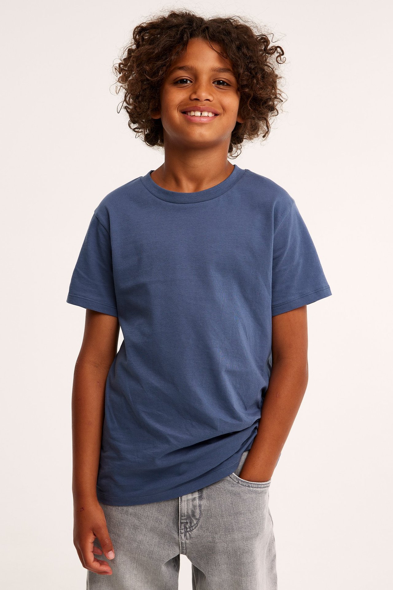 Widok z przodu uśmiechniętego dziecka w granatowym bawełnianym T-shircie i szarych dżinsach.