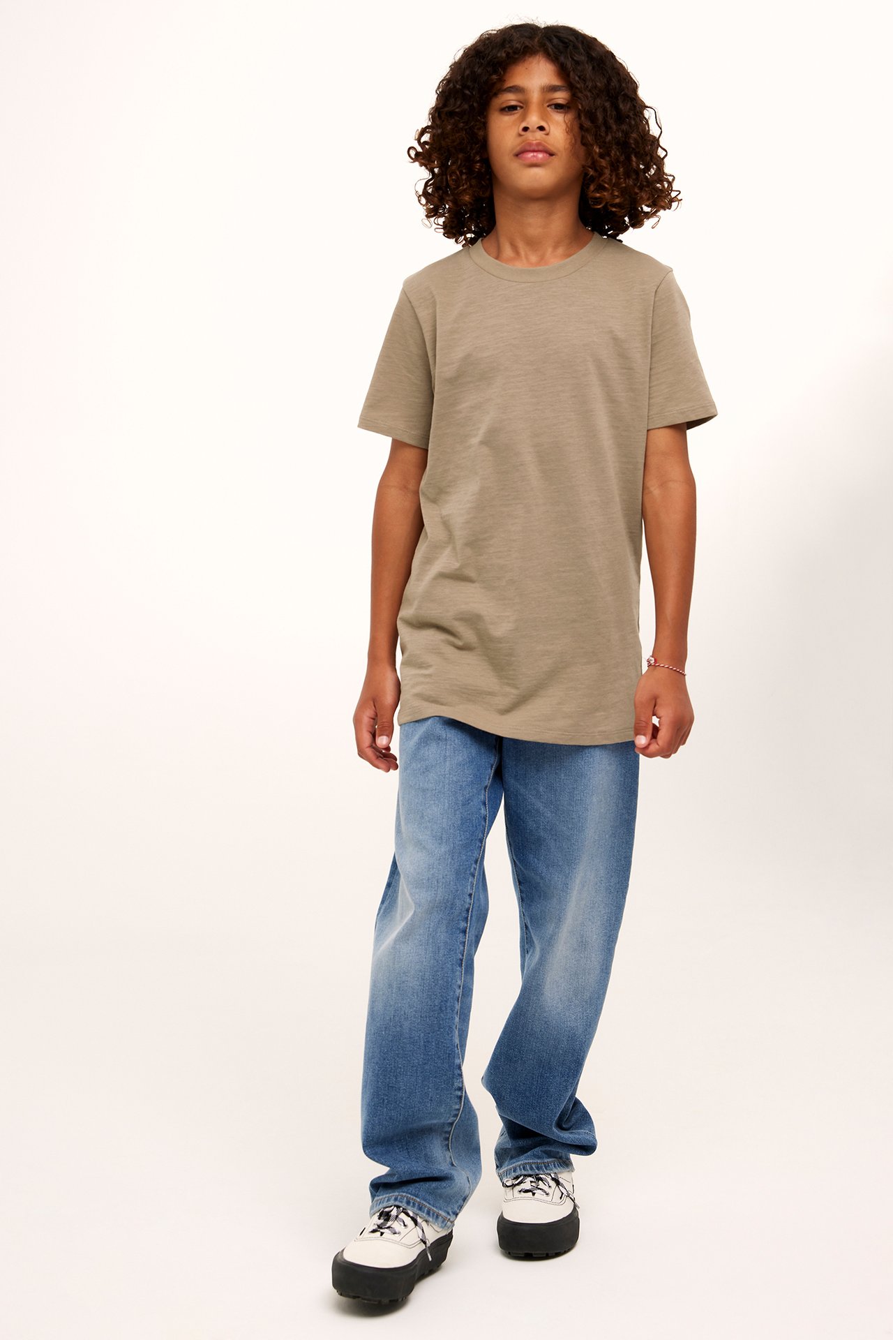 Framifrån: ett barn i enfärgad brun bomulls-t-shirt, lösa blå jeans och vita platåsneakers.