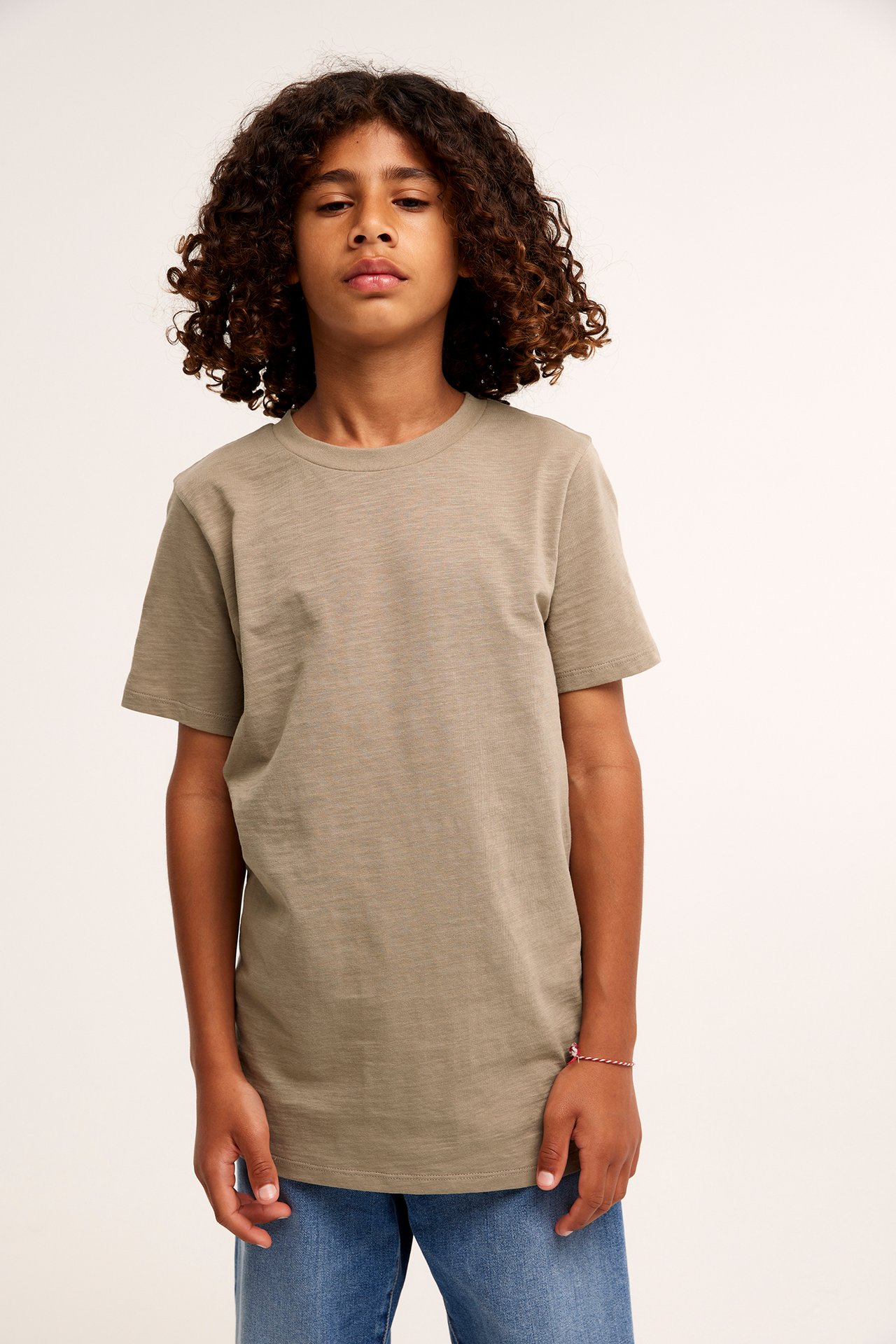 Frontvy av ett barn i enfärgad brun bomulls-t-shirt, blå jeans och ett rött och vitt armband.