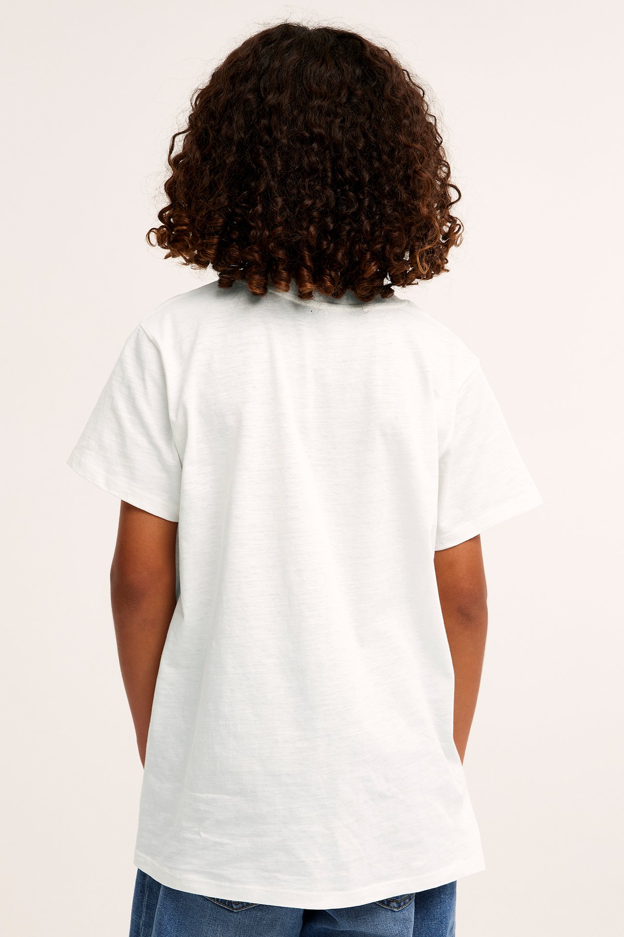 Bakifrån: ett barn i en offwhite kortärmad t-shirt i bomull och blå jeans.
