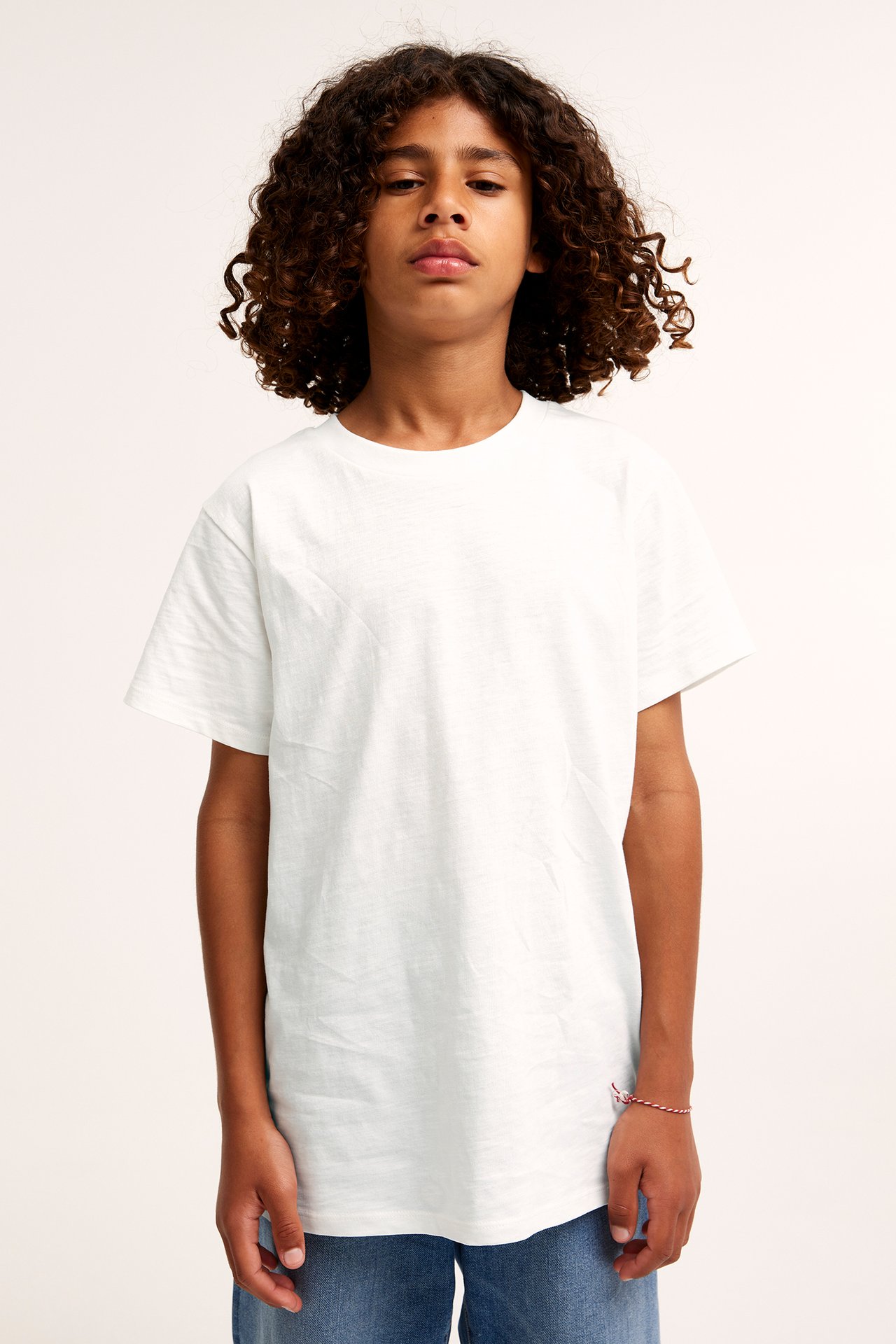 Framifrån: ett barn i offwhite kortärmad bomulls-t-shirt, blå jeans och randigt armband.