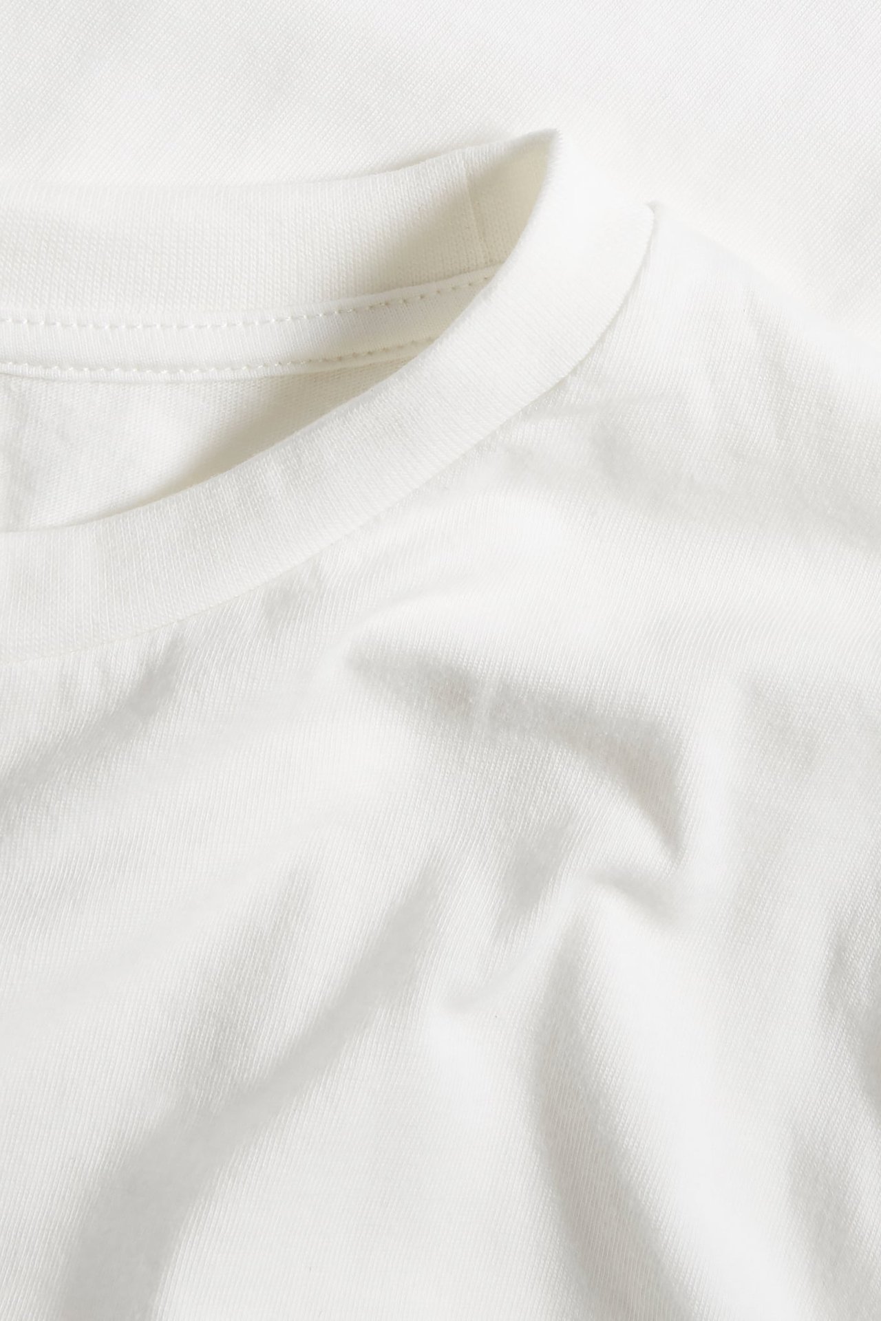 Zbliżenie na bawełniany T-shirt w kolorze ecru z prążkowanym okrągłym dekoltem i miękką fakturą materiału.