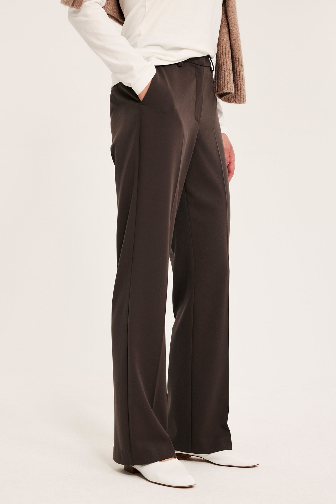 Sivukuva naisesta tummanruskeissa levenevissä housuissa, valkoisessa pitkähihaisessa paidassa ja vaaleanruskeassa neuleessa.