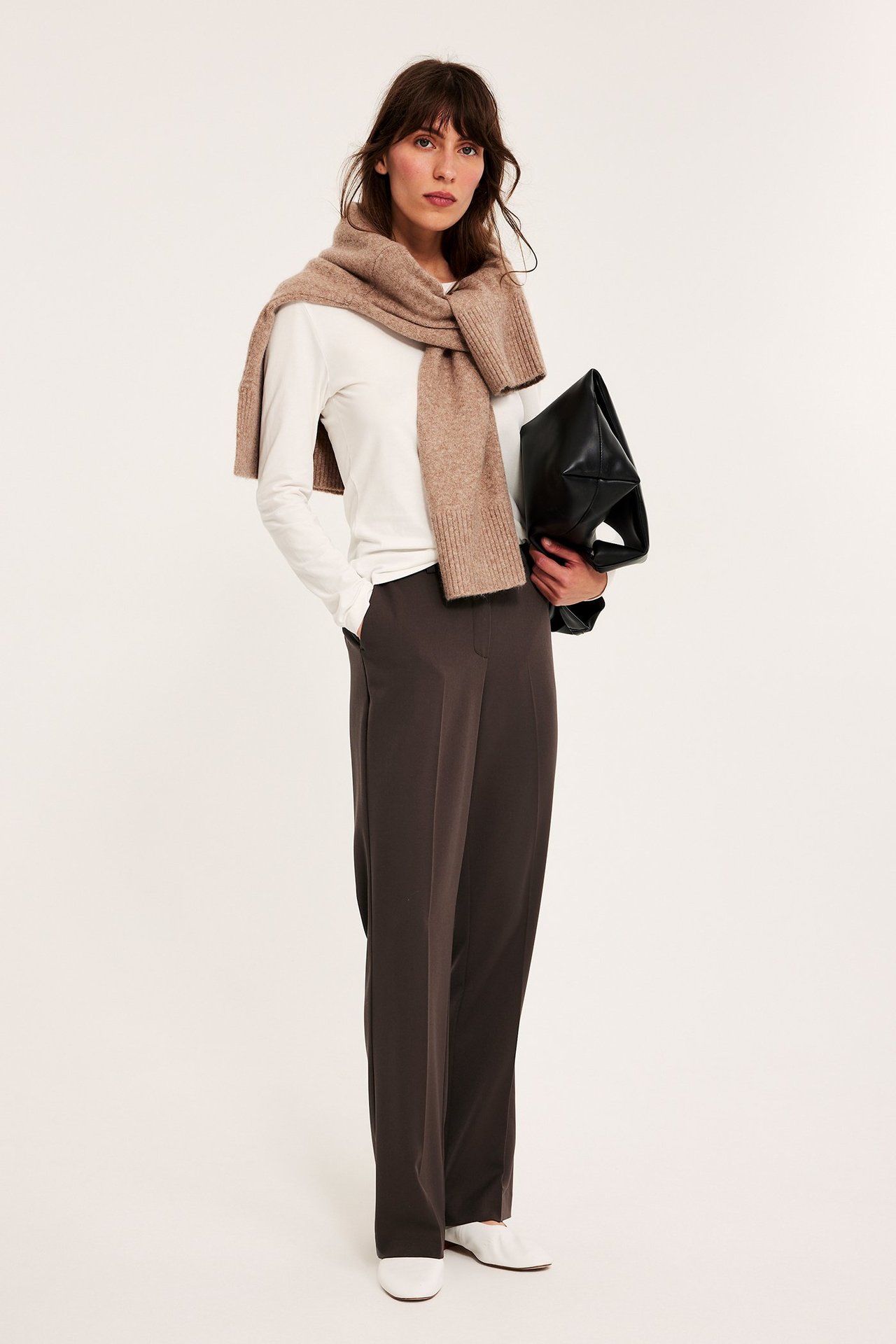 Edestä: Nainen tummanruskeissa suorissa housuissa, valkoinen toppi, beige neulottu huivi, pitää mustaa laukkua.