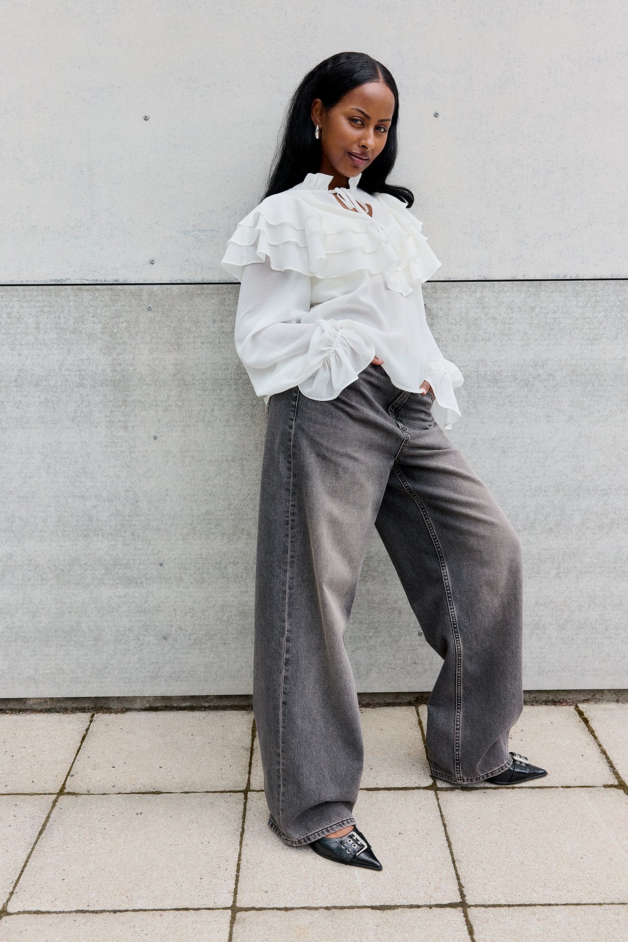 Framifrån: kvinna i tvättade svarta baggy jeans, vit volangblus och svarta spetsiga skor.