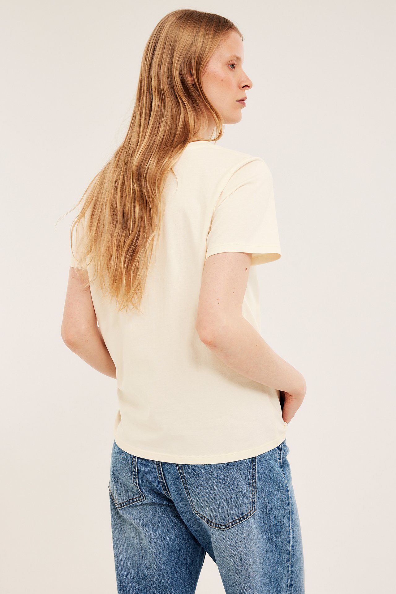 Naisen selkäpuoli offwhite-puuvilla-T-paidassa, jossa on suora istuvuus, ja sinisissä farkuissa.