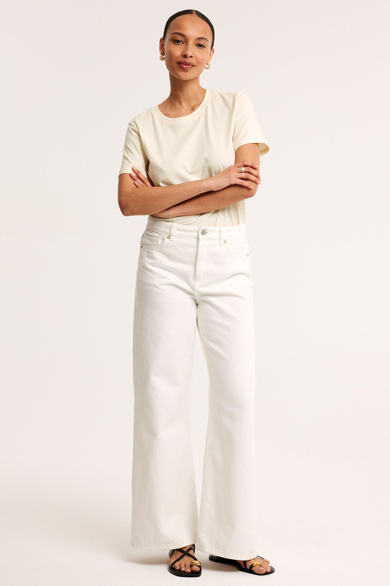 Frontbilde av en kvinne i en offwhite rett bomulls-T-skjorte, hvite vide jeans og svarte sandaler.