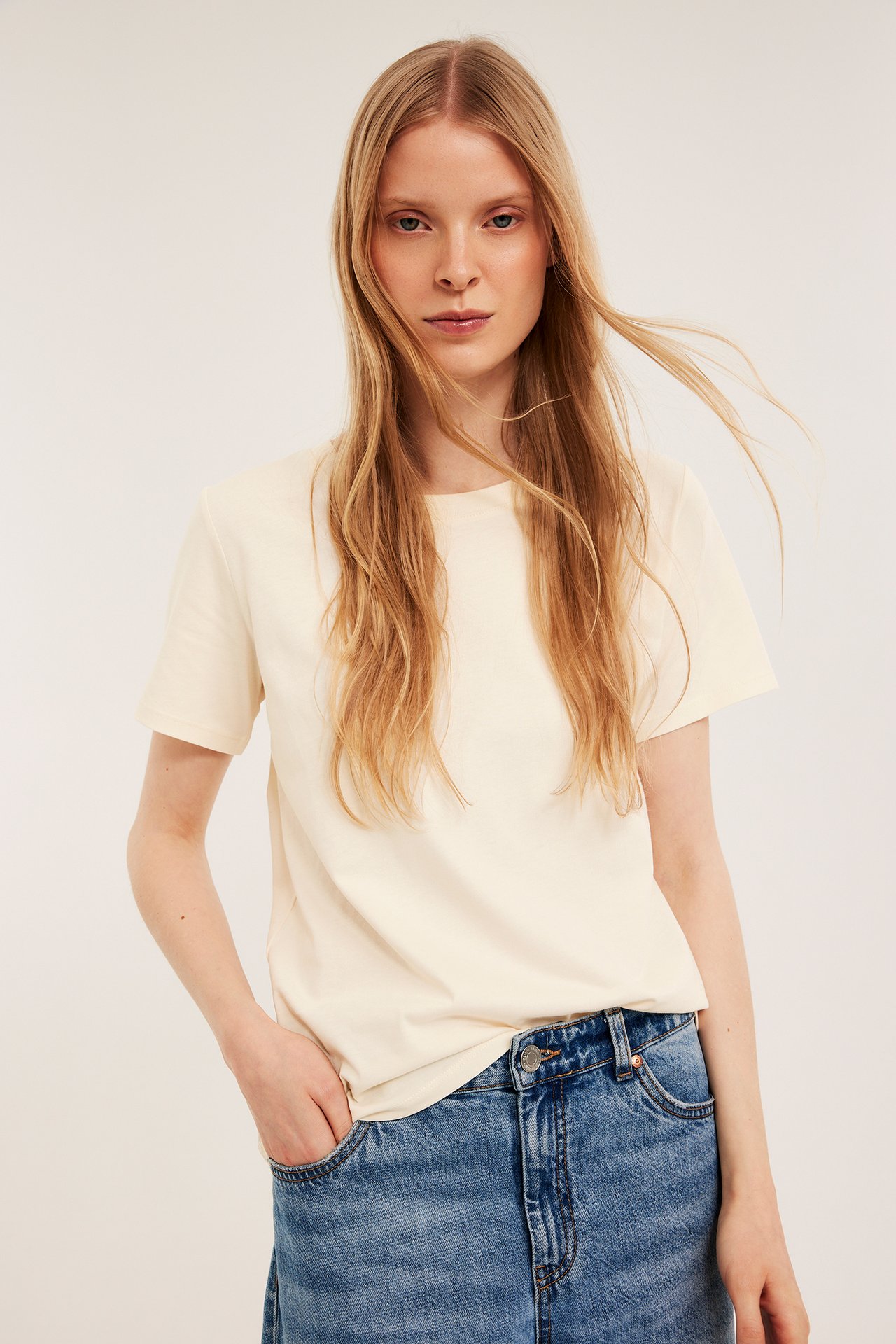 Frontbilde av kvinne i en offwhite, rett bomulls-T-skjorte og blå jeans med hånden i lommen.