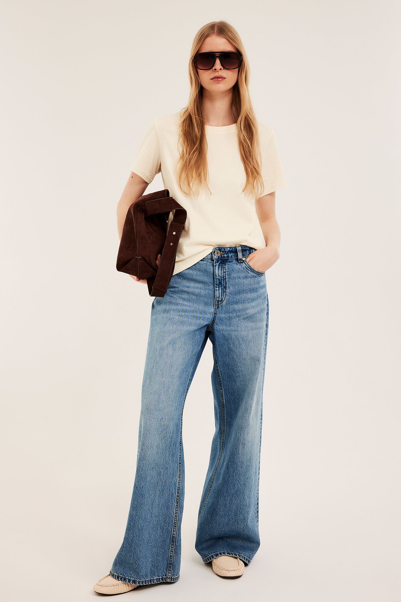 Nainen edestäpäin: luonnonvalkoinen T-paita, siniset farkut, beiget mokkasiinit, tummat aurinkolasit, ruskea laukku kädessä.