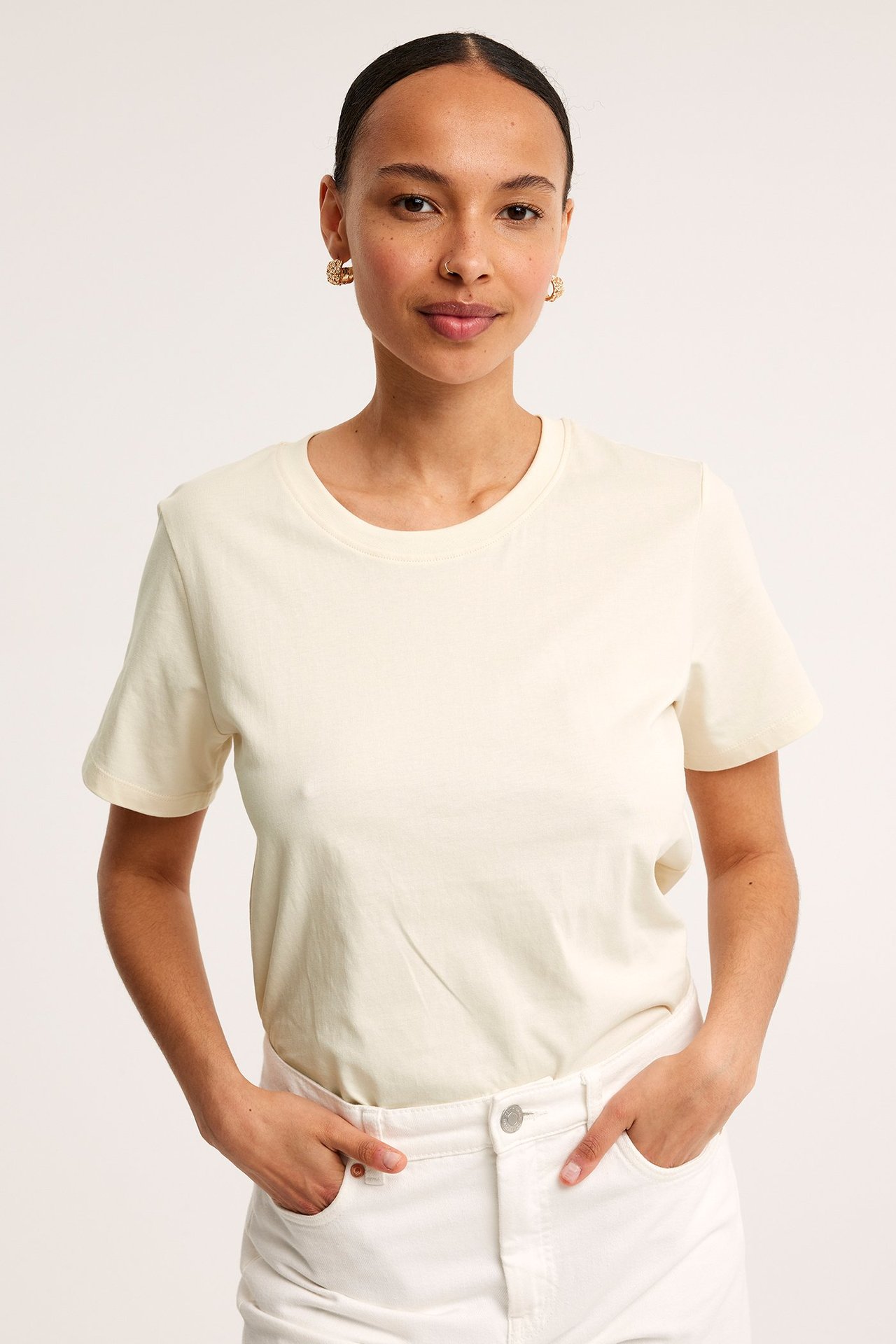 Frontbilde av en kvinne i en offwhite, rett bomulls-T-skjorte stukket ned i hvite jeans, med gulløreringer.