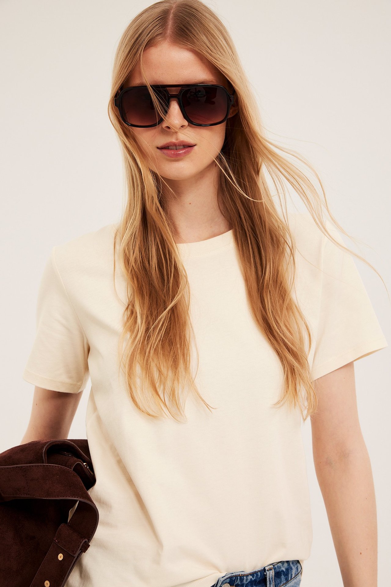 Närbild på kvinna i offwhite bomulls-T-shirt och mörka solglasögon. Hon håller en brun jacka.