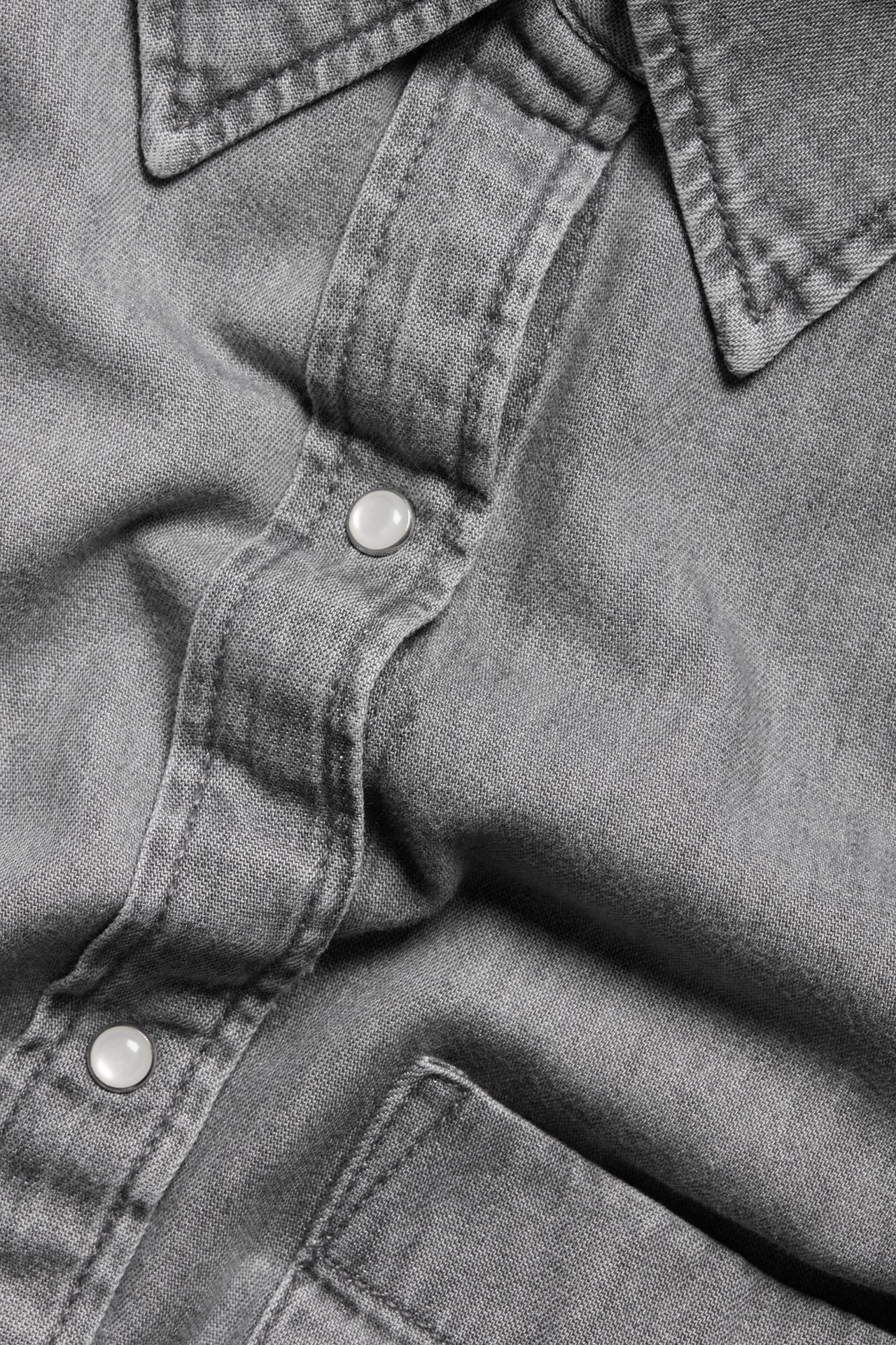 Nærbilde av en grå denimskjortekjole med krage, hvite trykknapper og en brystlomme.