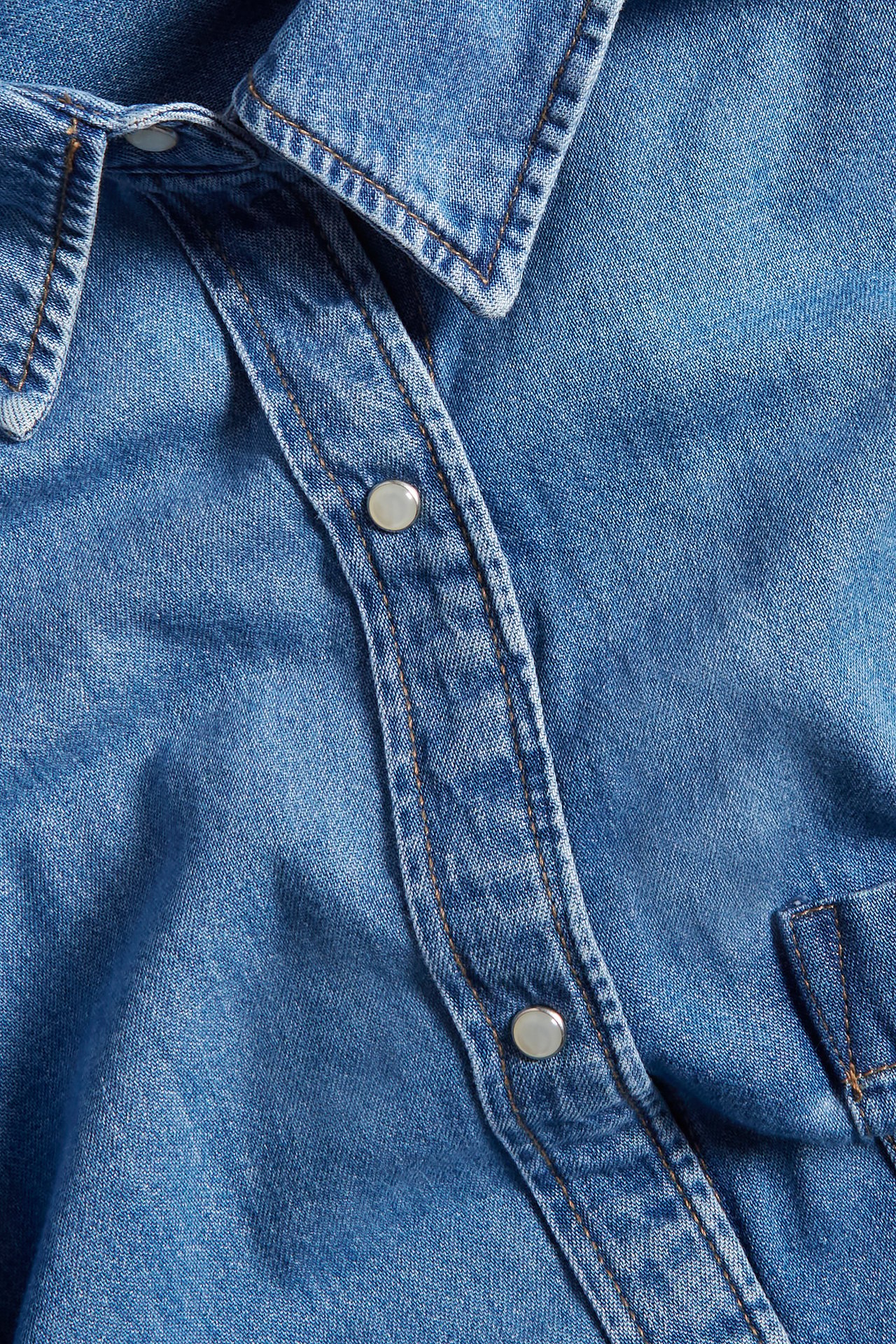 Nærbilde av en lyseblå denimskjortekjole med krage, sølvfargede trykknapper og oransje sømmer.