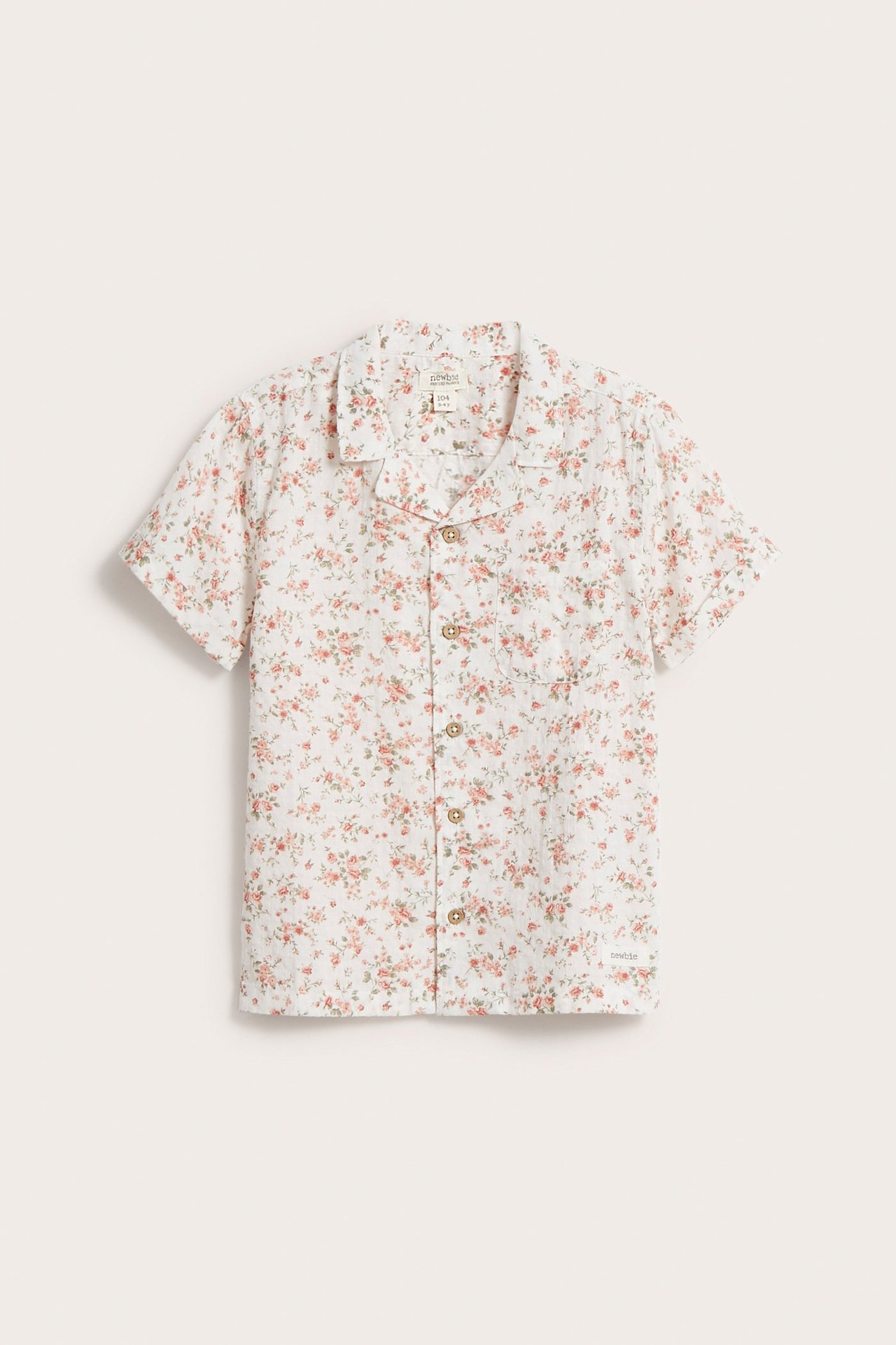 Framifrån: Vit kortärmad barnskjorta i bomull med rosa blommönster, slagkrage, ficka och knappar.