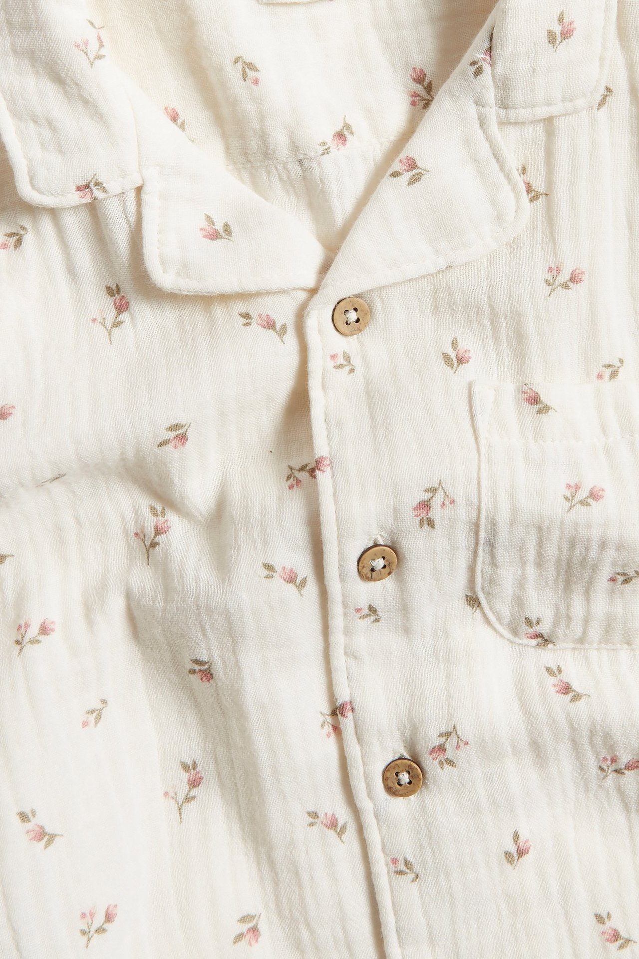 Närbild framifrån: beige kortärmad bomullsskjorta med små rosa blommönster, krage, träknappar, bröstficka.