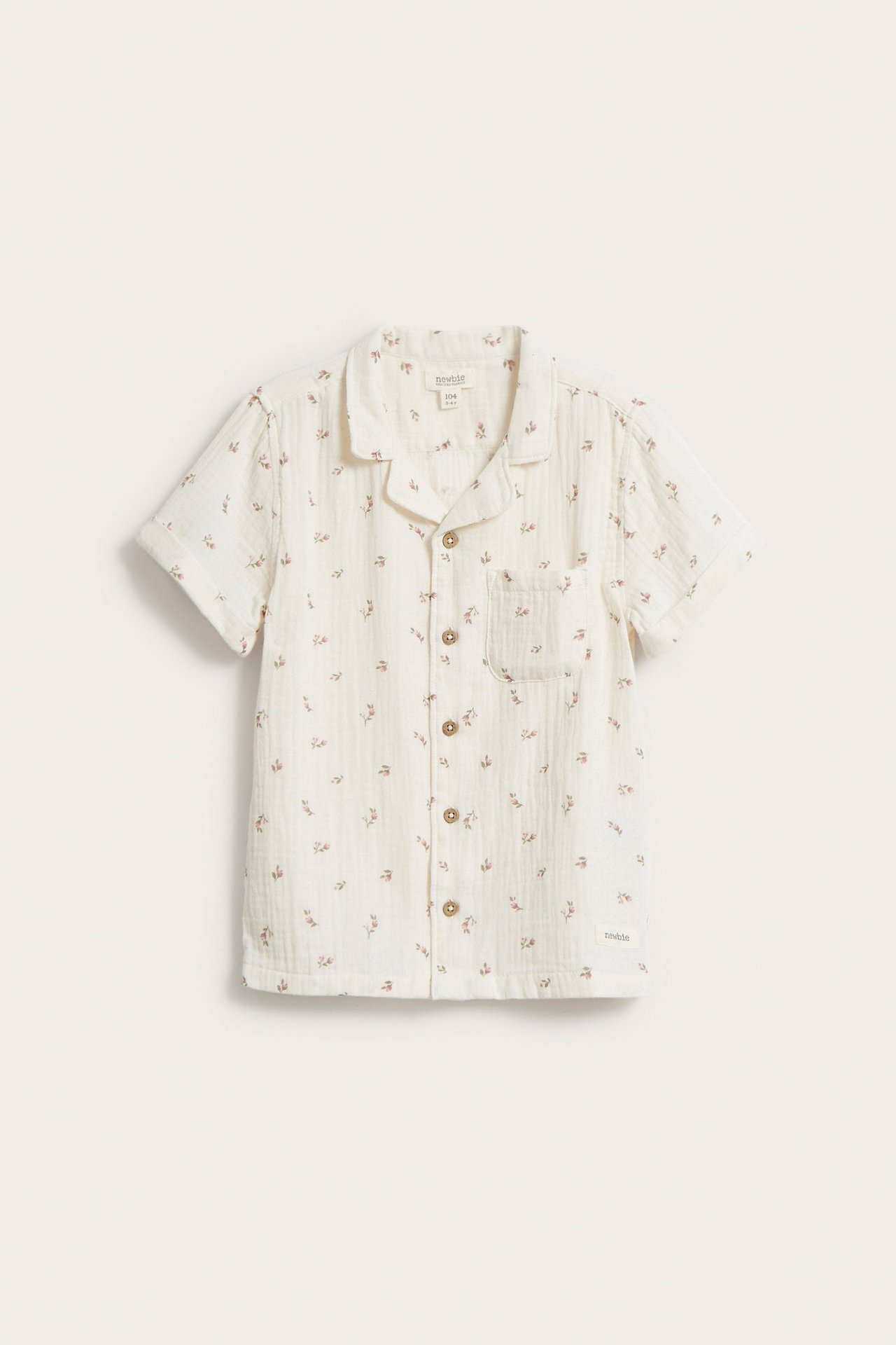 Framifrån, en beige blommig kortärmad skjorta i bomullsmuslin för barn, med krage och knappar.