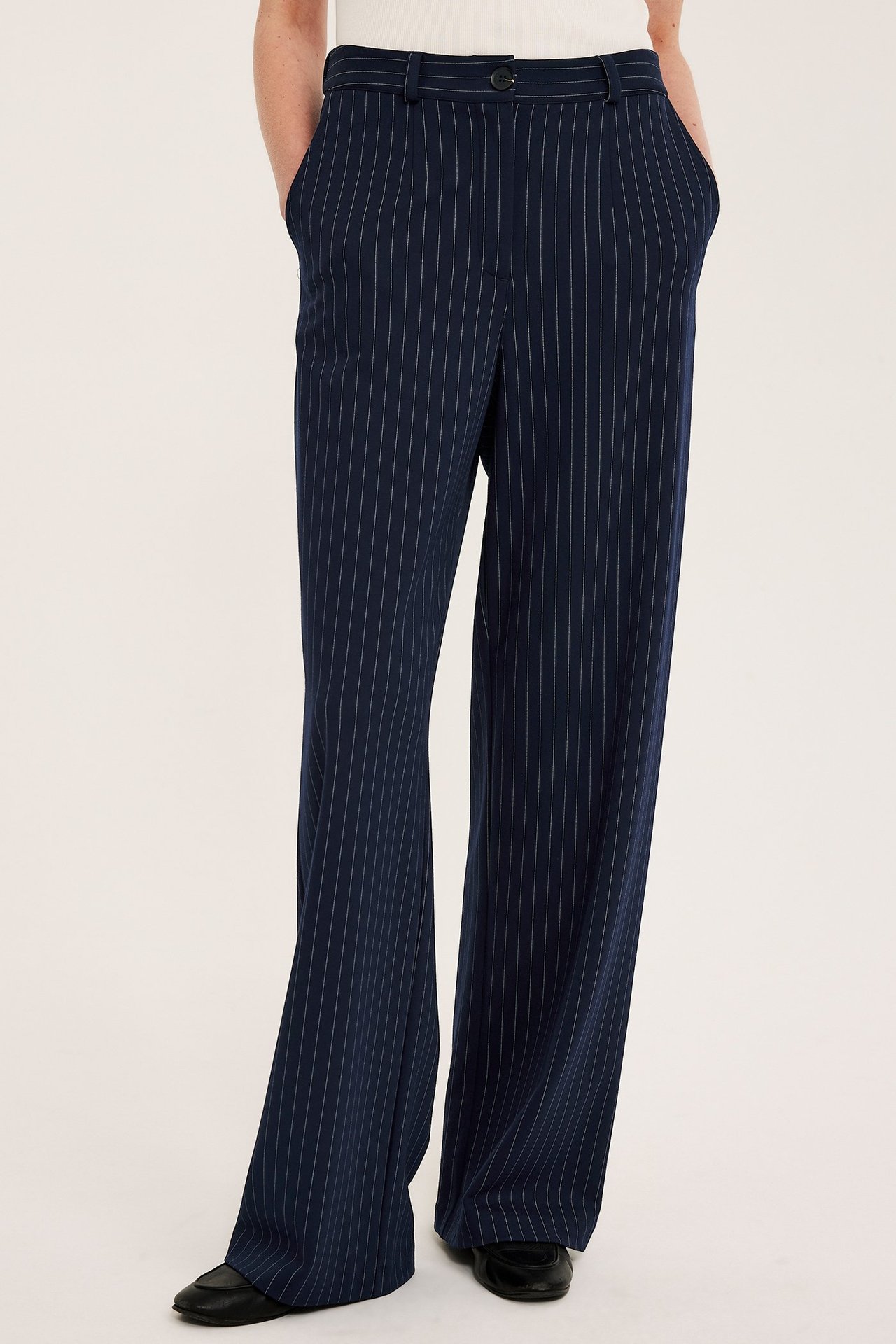 Widok z przodu: kobieta w granatowych spodniach garniturowych w paski, białym topie i czarnych butach.