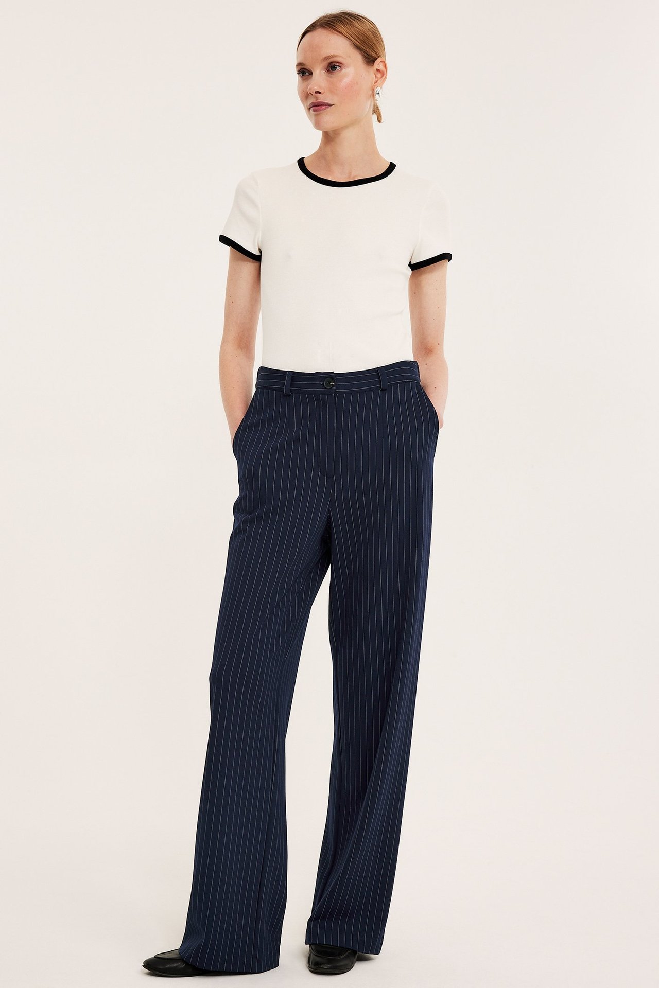 Edestäpäin, nainen tummansinisissä raidallisissa puvunhousuissa ja luonnonvalkoisessa t-paidassa mustalla reunalla.