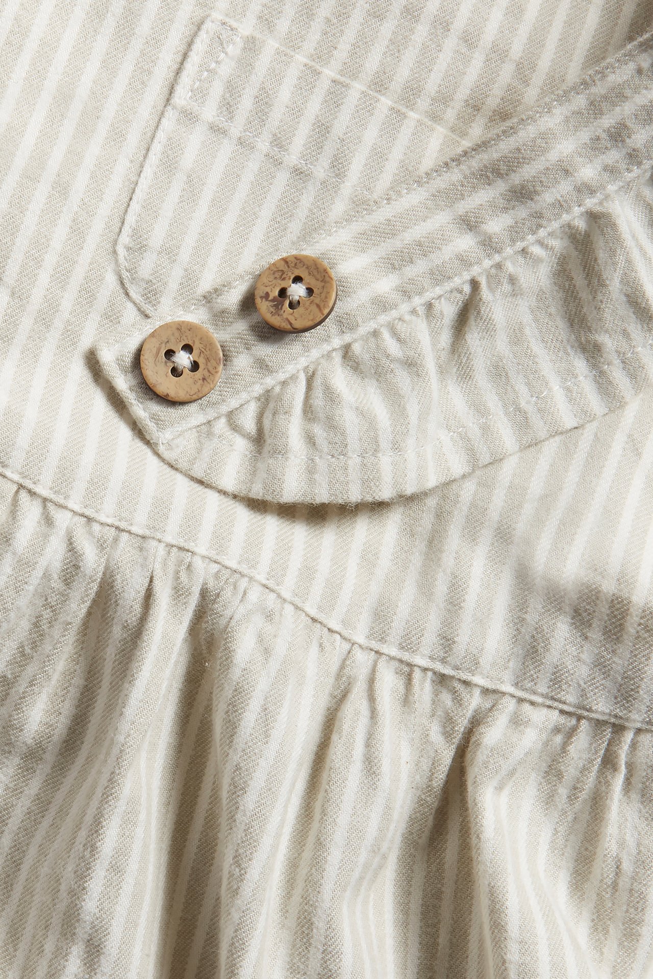 Närbild av en randig beige och benvit hängselklänning med två träknappar och en liten bröstficka.