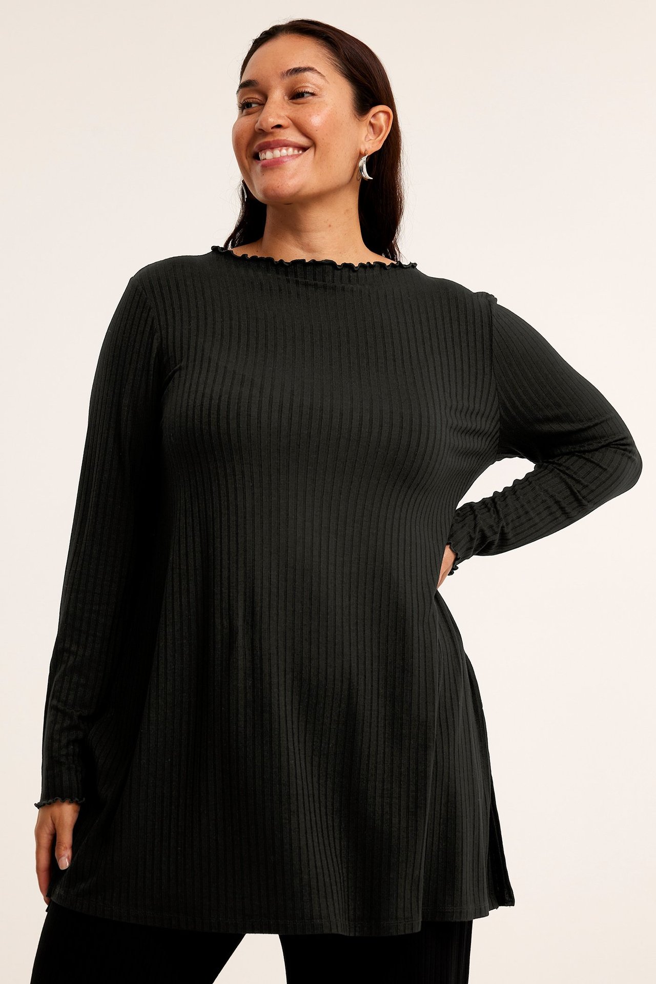 Widok z przodu uśmiechniętej kobiety w czarnej prążkowanej tunice z falbankami, czarnych spodniach i srebrnych kolczykach.