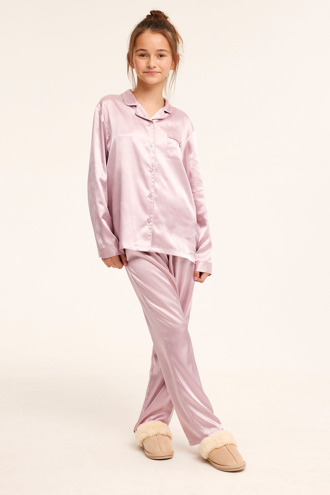 Framifrån, ett barn i rosa satinpyjamas med knäppt överdel och lösa byxor, samt beiga fluffiga tofflor.