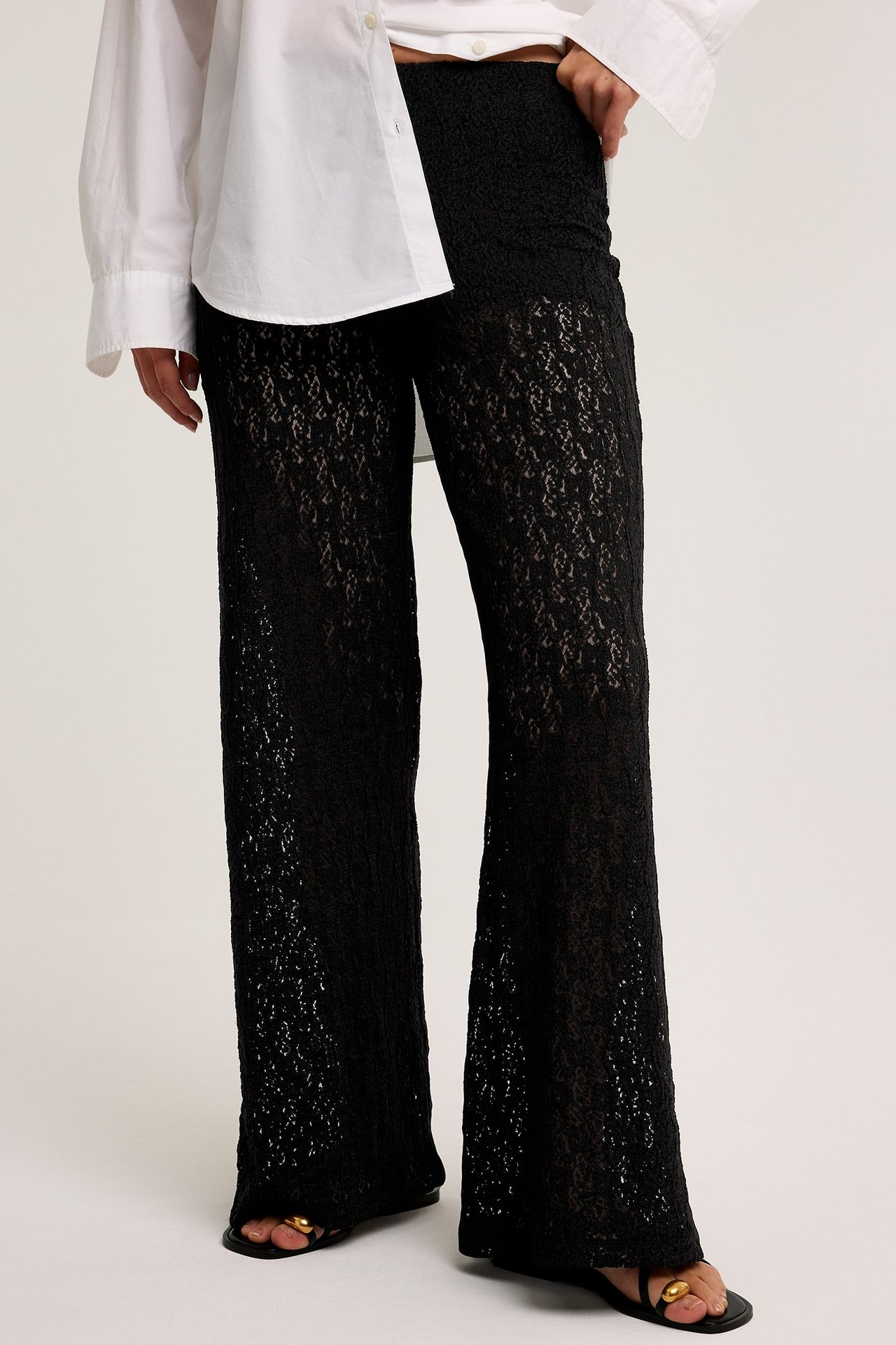 Framifrån: kvinna i svarta vida spetsbyxor, vit oversized skjorta och svarta platta sandaler.