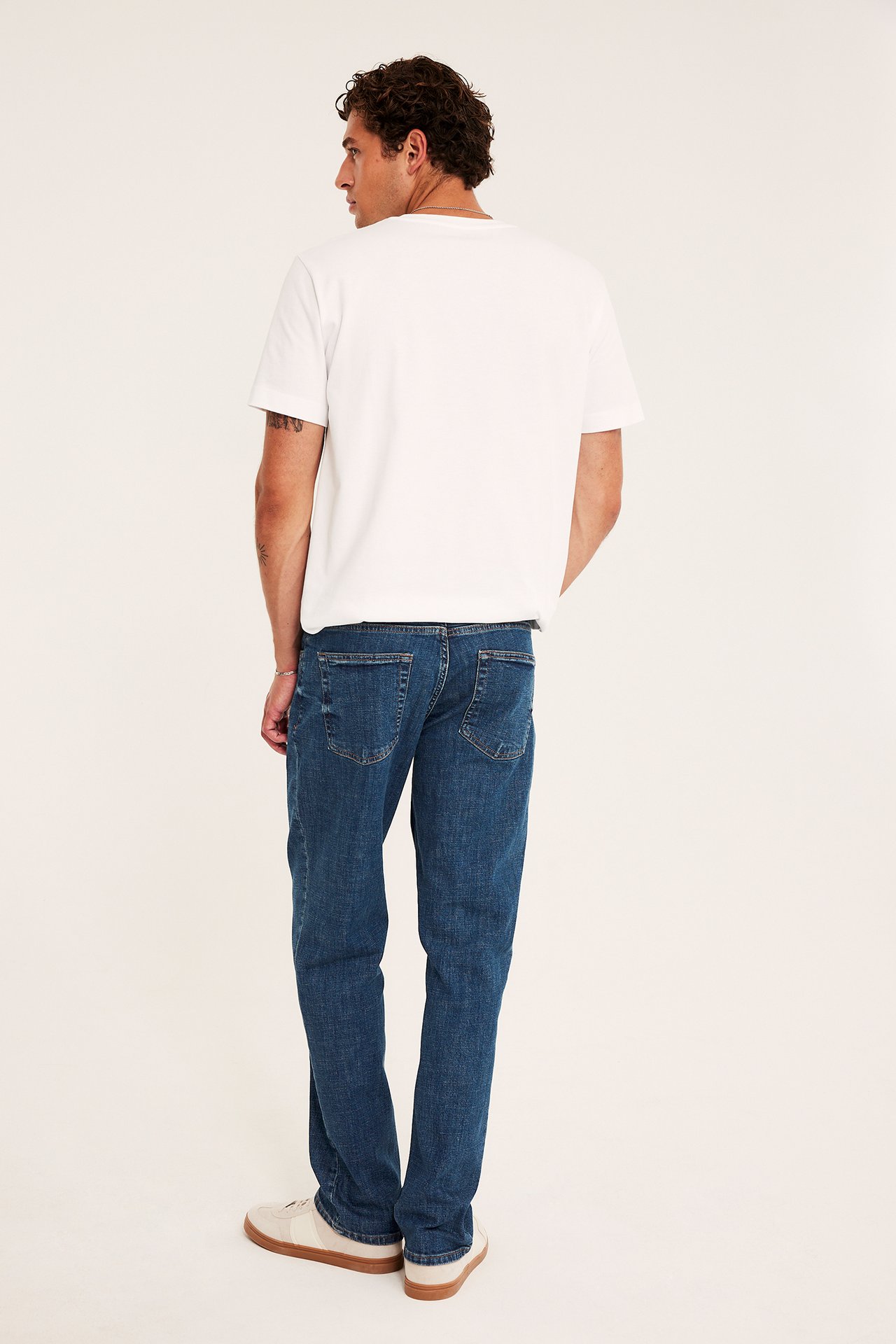Ryggvisning av mann i hvit kortermet t-skjorte, blå jeans og beige joggesko.