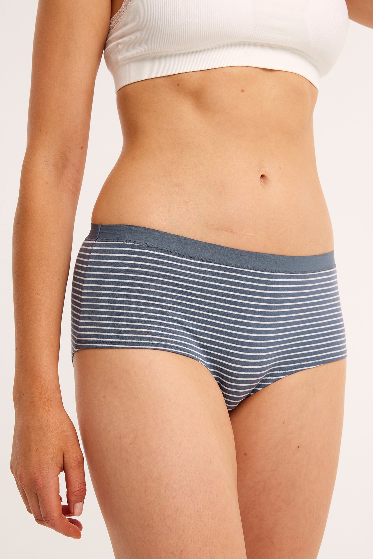 Framifrån, kvinna i blågrå randiga bomullshipstertrosor och en vit ribbad bralette.