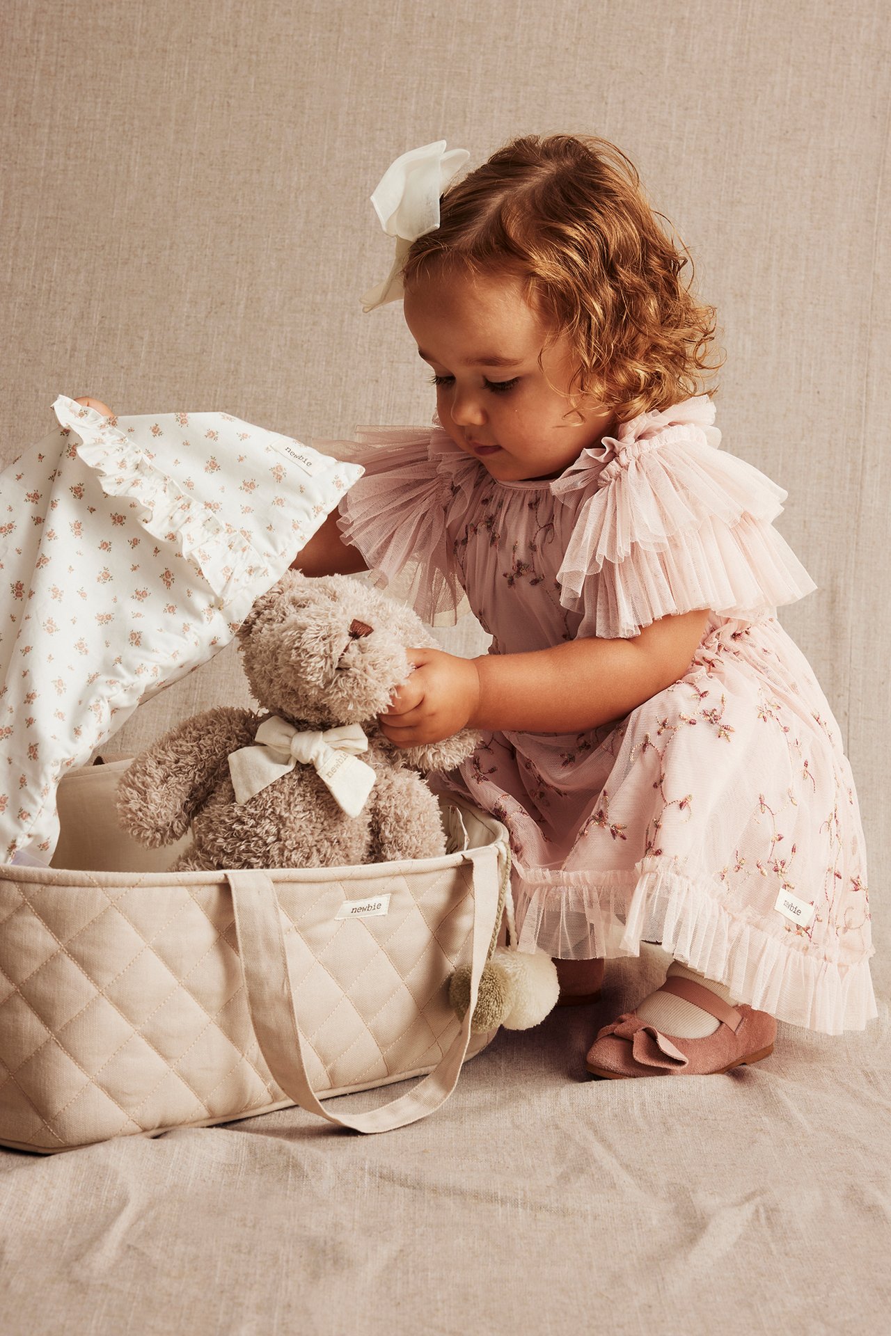 Baby i lys rosa blomstret nettingkjole, rosa sko, holder teddybjørn og blomstret teppe i vattert veske.