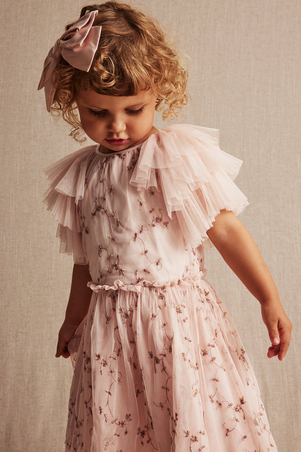 Frontvisning: baby i en lyserosa blomstret brodert nettingkjole med volangermer og en rosa hårsløyfe.