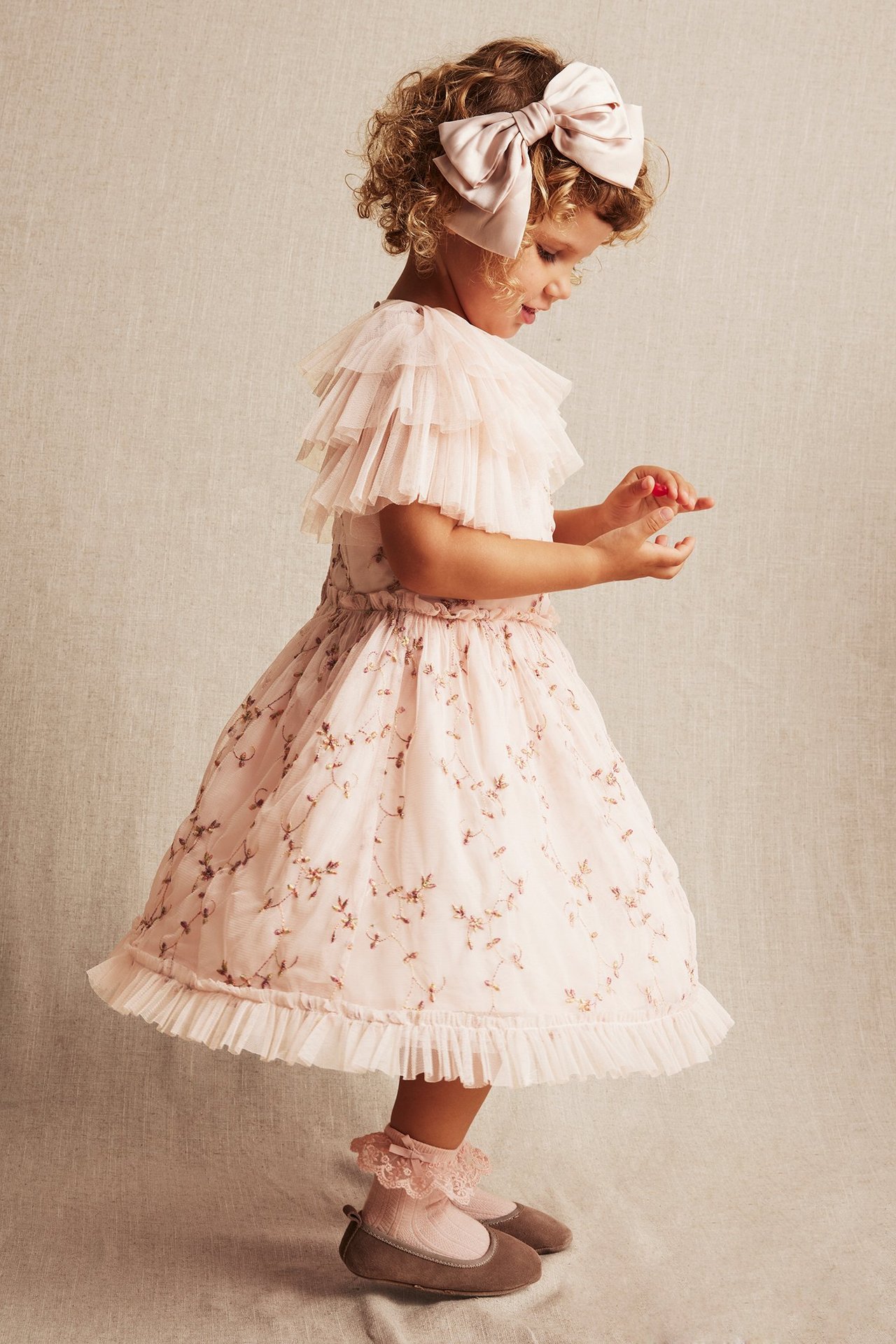 Widok z boku dziecka w jasnoróżowej haftowanej sukience, różowej kokardzie, różowych skarpetkach i brązowych baletkach.