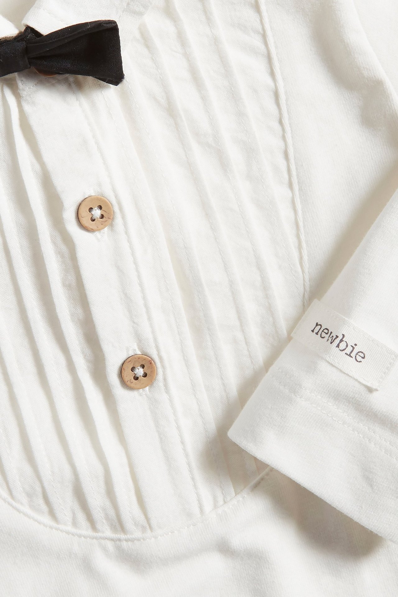 Zbliżenie na ecru body smokingowe dla niemowląt z plisami z przodu, drewnianymi guzikami i czarną muchą.