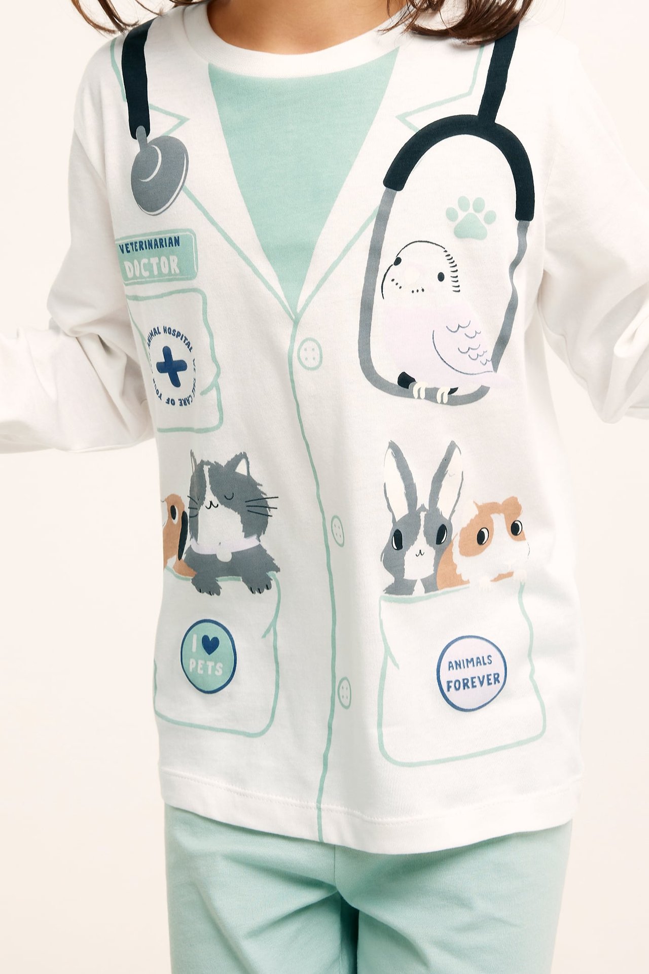 Framifrån: ett barn i vit bomullspyjamas med veterinärtryck och ljus turkosa pyjamasbyxor.
