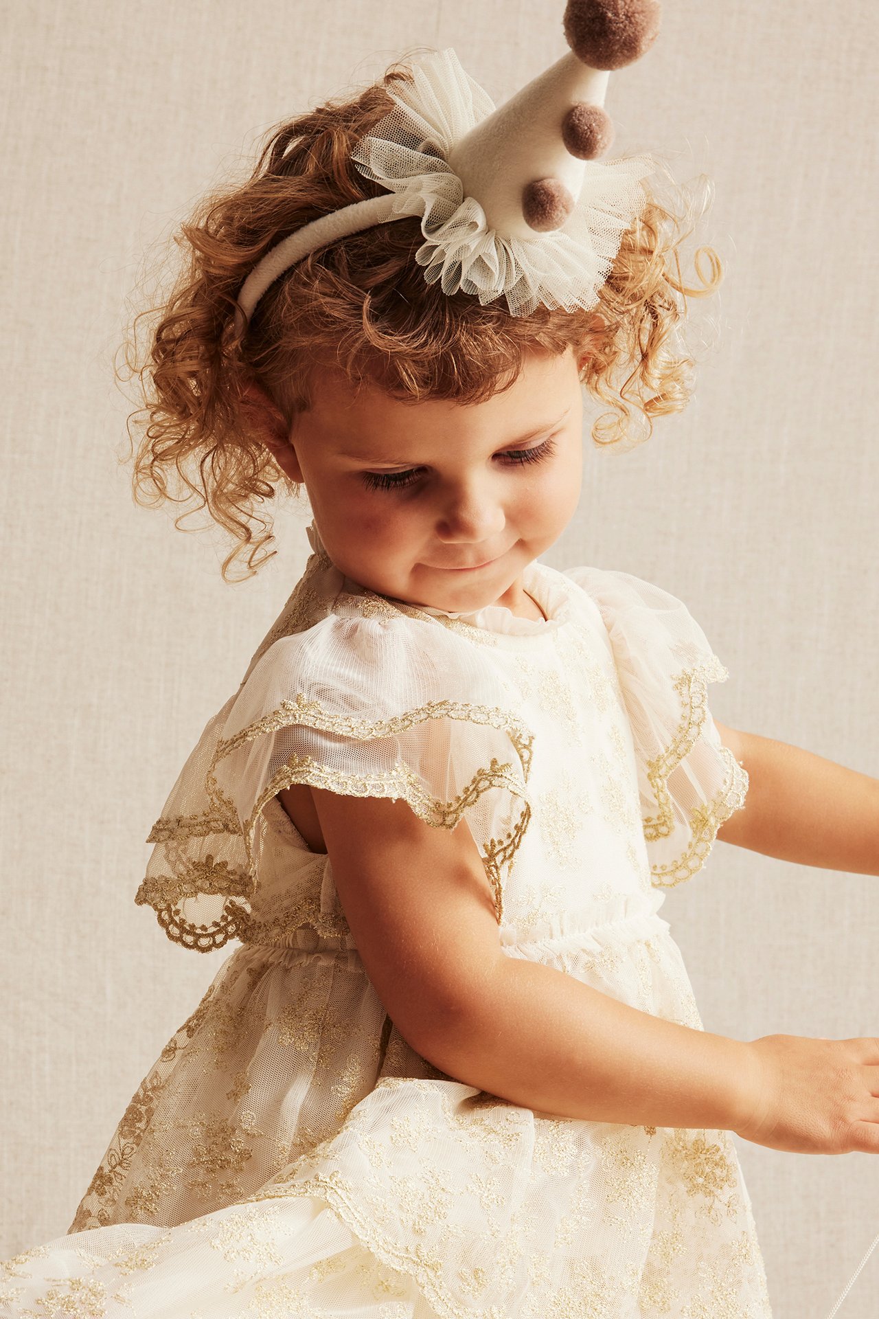 Närbild på ett barn i en offwhite broderad meshklänning med gulddetaljer och ett festhatt-diadem.