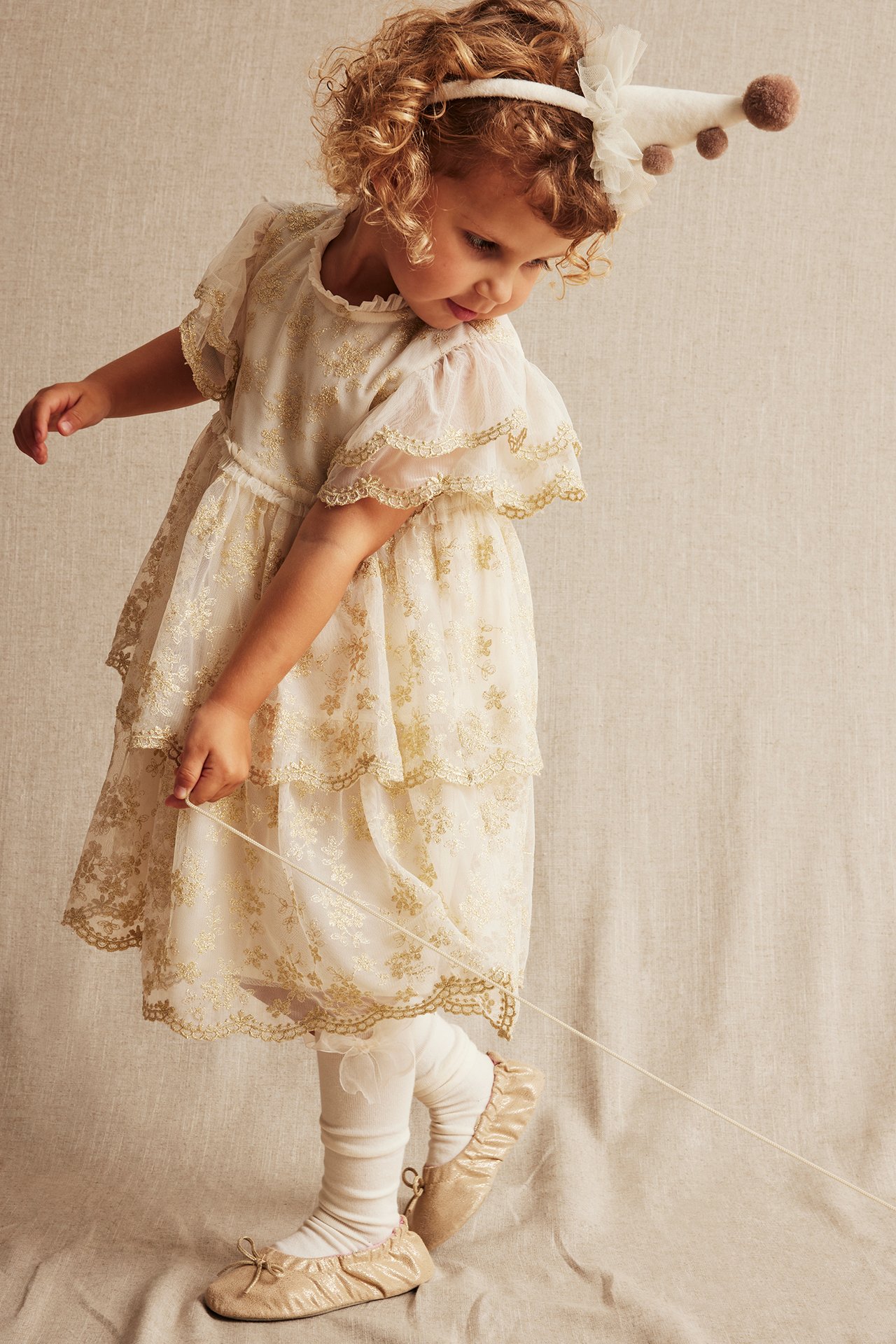Framsida av ett barn i en offwhite broderad meshklänning, vita strumpbyxor, beiga ballerinaskor och en festhatt.