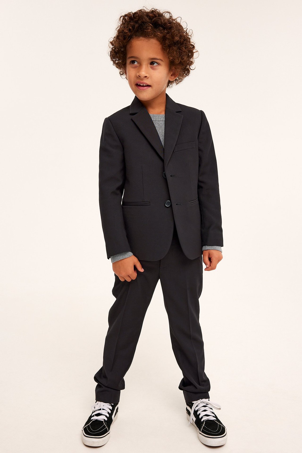 Framifrån, ett barn i svart enkelknäppt kavaj, svarta byxor, grå tröja och svarta sneakers.