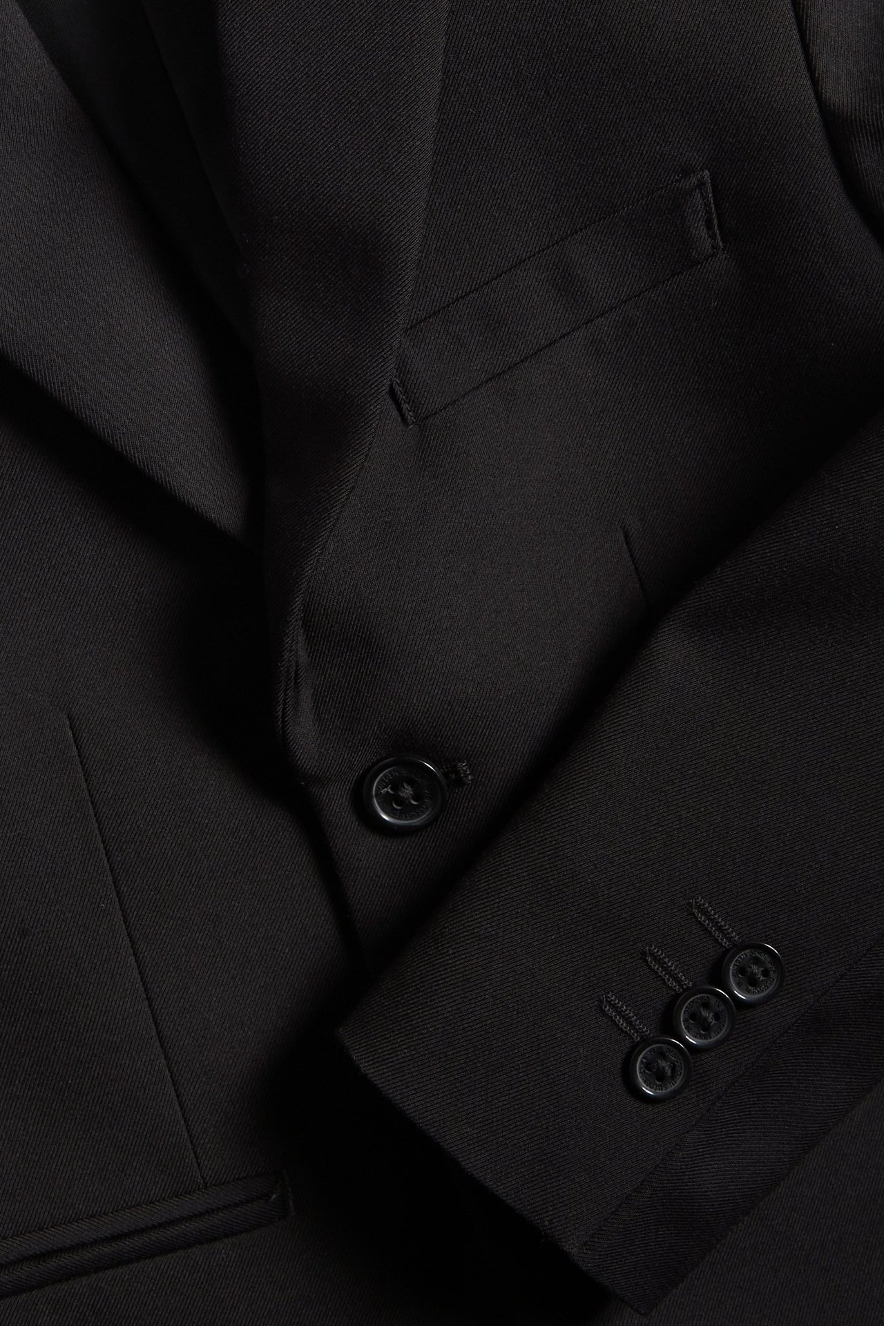 Zbliżenie czarnej marynarki dziecięcej z jednym guzikiem, kieszenią z wypustką i trzema guzikami na mankiecie.