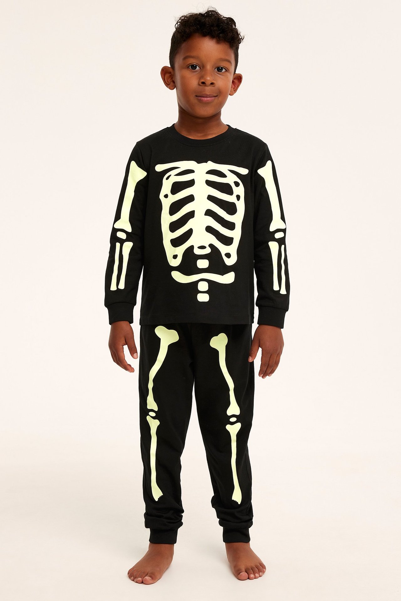 Edestä kuvattu lapsi mustassa puuvillapyjamassa, jossa pimeässä hohtava luurankoprintti.