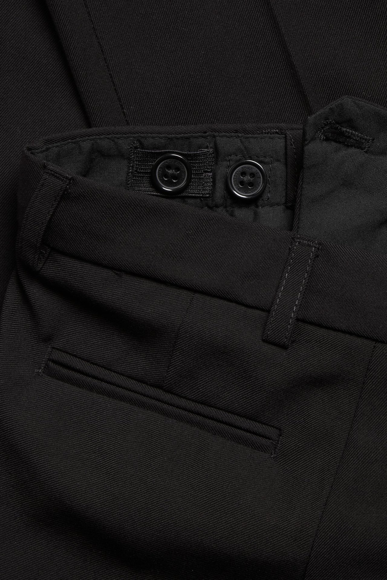 Zbliżenie czarnych spodni garniturowych, pokazujące regulowaną talię z guzikami i kieszeń z wypustką.