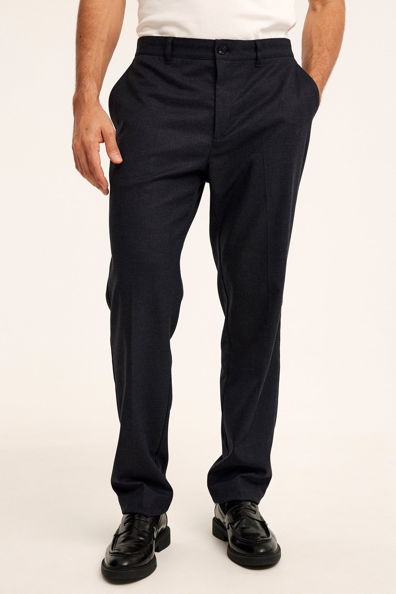 Edestä kuvattu mies yllään tummanharmaat meleeratut puvunhousut prässivekeillä, valkoinen t-paita ja mustat loaferit.