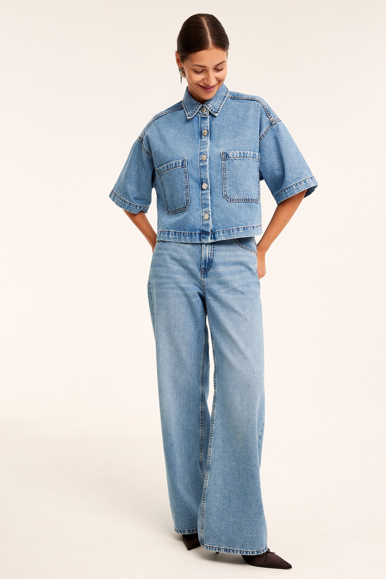 Framifrån: Kvinna i kortärmad ljusblå jeansskjorta, vida jeans och svarta spetsiga skor.