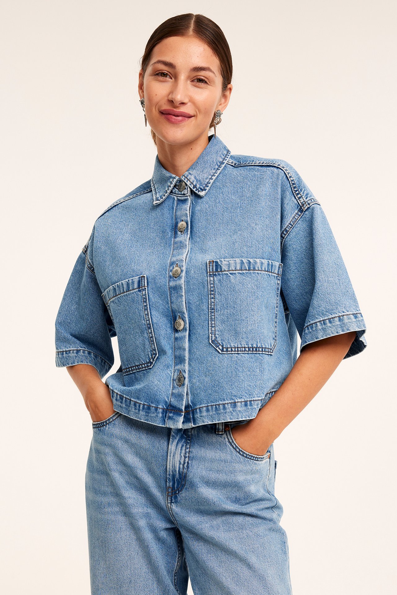 Framifrån: Kvinna i ljusblå kortärmad jeansskjorta med knappar, fickor och matchande jeans.
