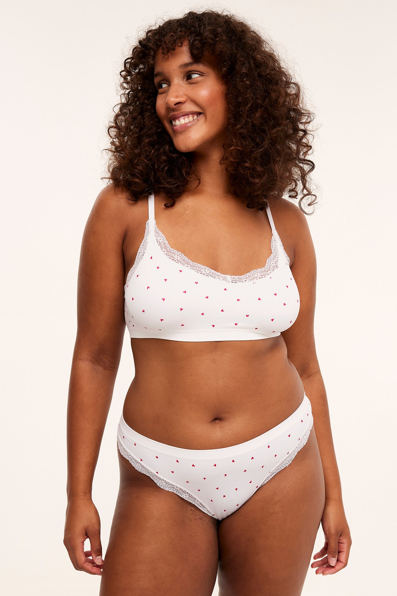 Framifrån, kvinna i vit bralette med röda hjärtan och spetskant, matchande trosor.