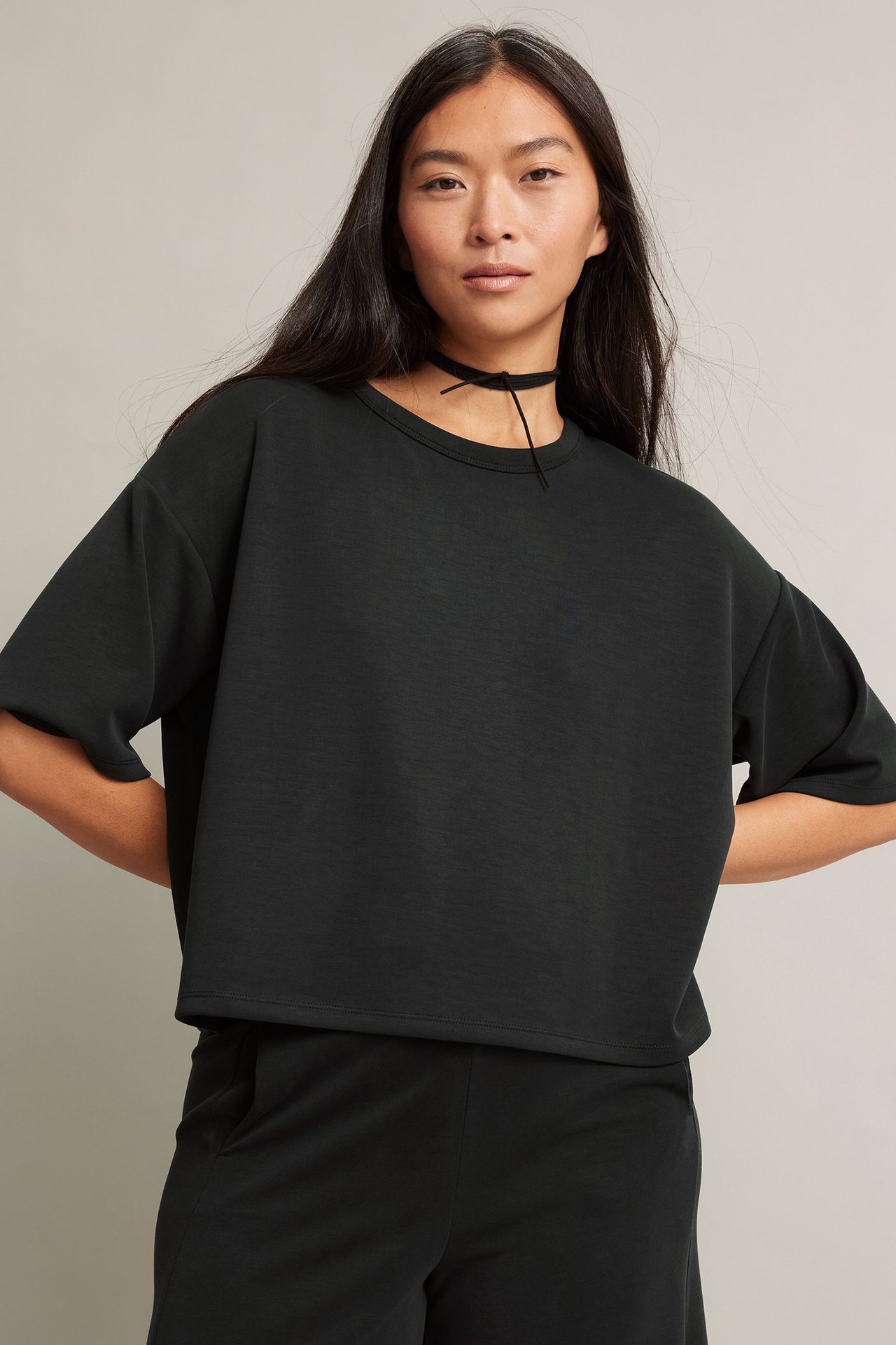 Framifrån, kvinna i svart, boxig T-shirt med längre ärmar, svarta byxor och ett chokerhalsband.