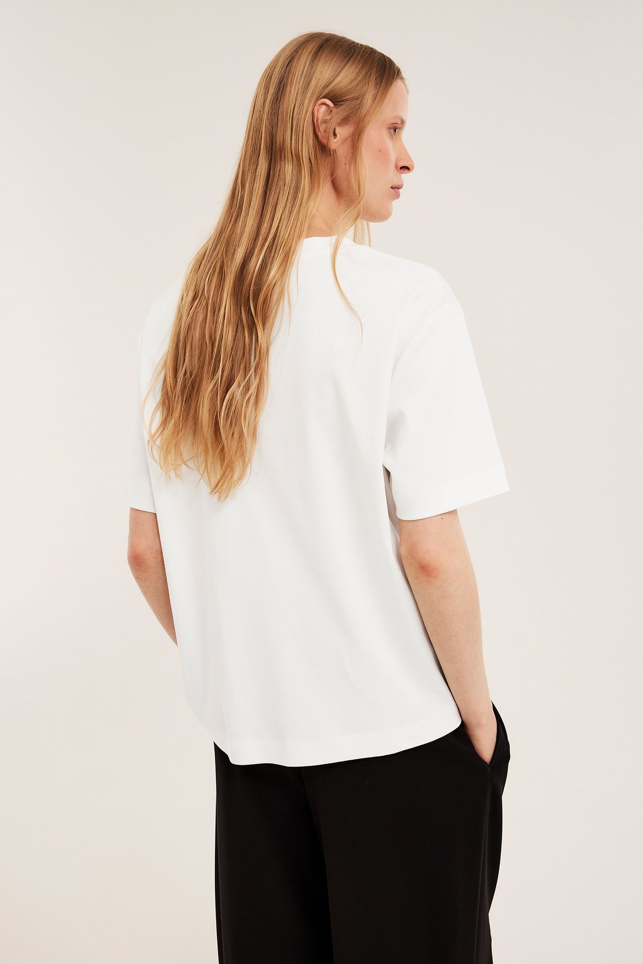 Naisen takakuva yllään luonnonvalkoinen oversized interlock-T-paita ja mustat leveälahkeiset housut.